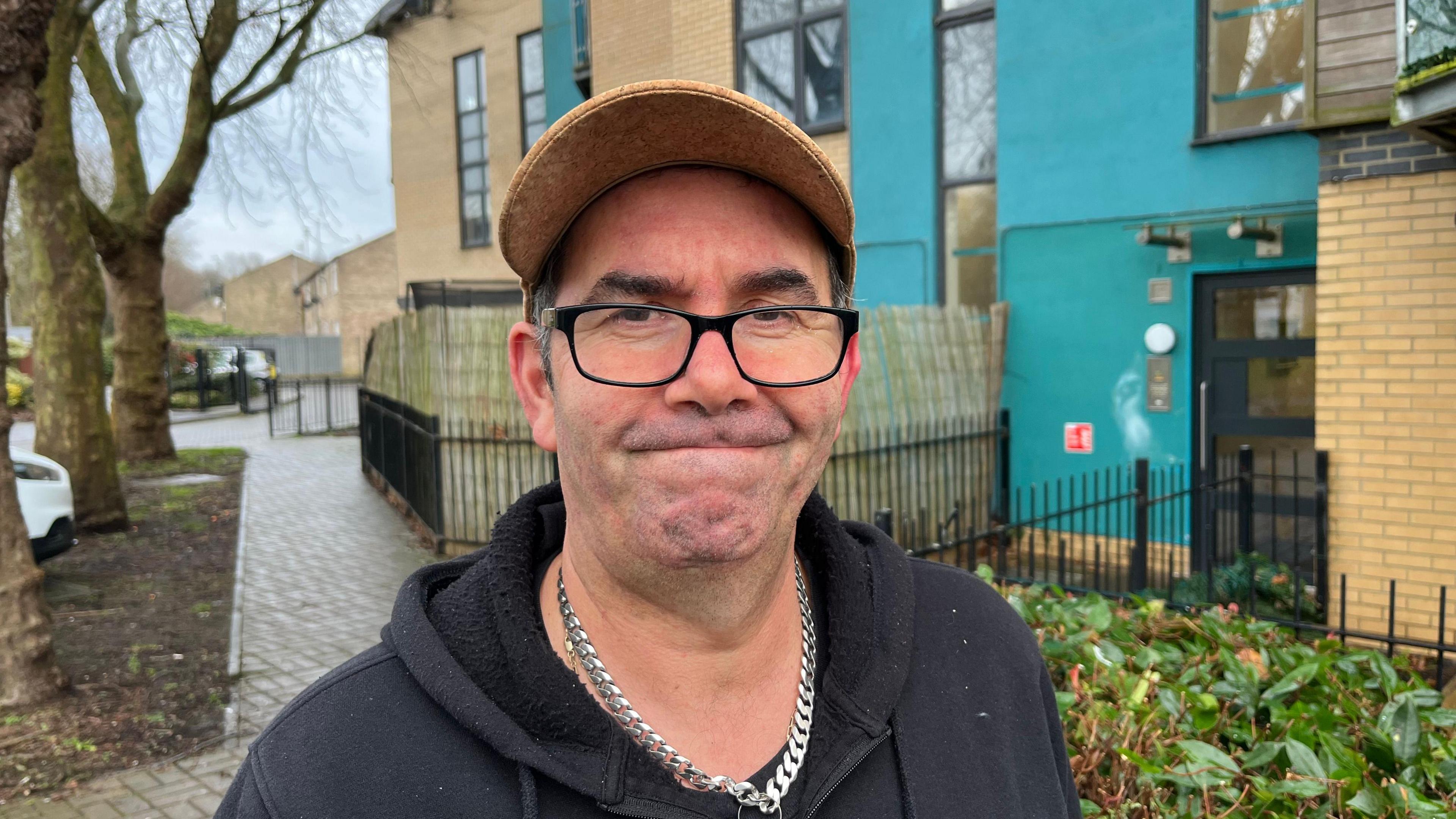 Nuno Bastos, is wearing a light brown peaked cap, black rimmed spectacles, he has short black/grey hair visible on either side of his head. He is wearing a dark blue hoodie and has a chunky silver linked chain around his neck, behind him are block-paved pathways in front of black metal railings the flat is brick and render, which is turquoise