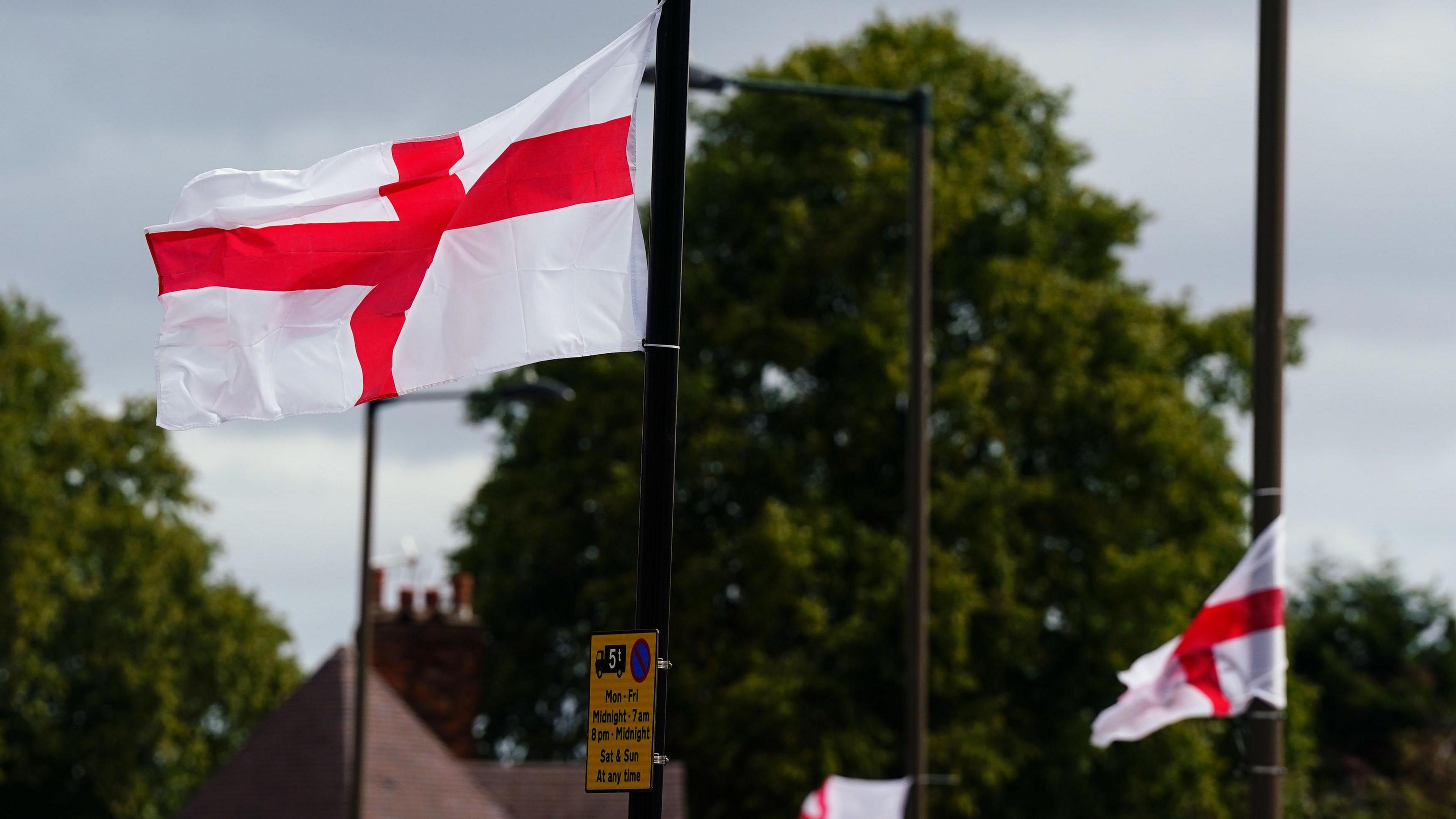 St George's flags fly from lamp-posts in Highters Heath, south Birmingham. We can see them blowing in the wind with the outline of trees and houses behind them. There is a yellow street sign off in the distance.