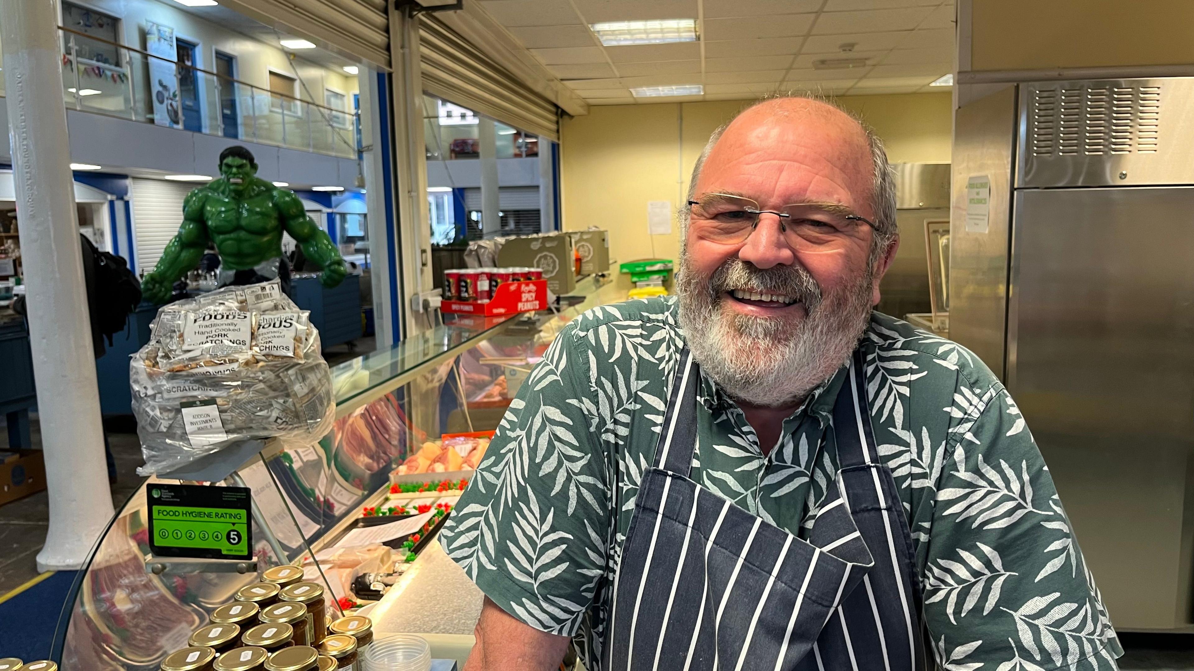 Stephen Holgate behind the counter at his stand in Pannier Market wearing a teal shirt with white leaves on it and a striped blue and white apron.