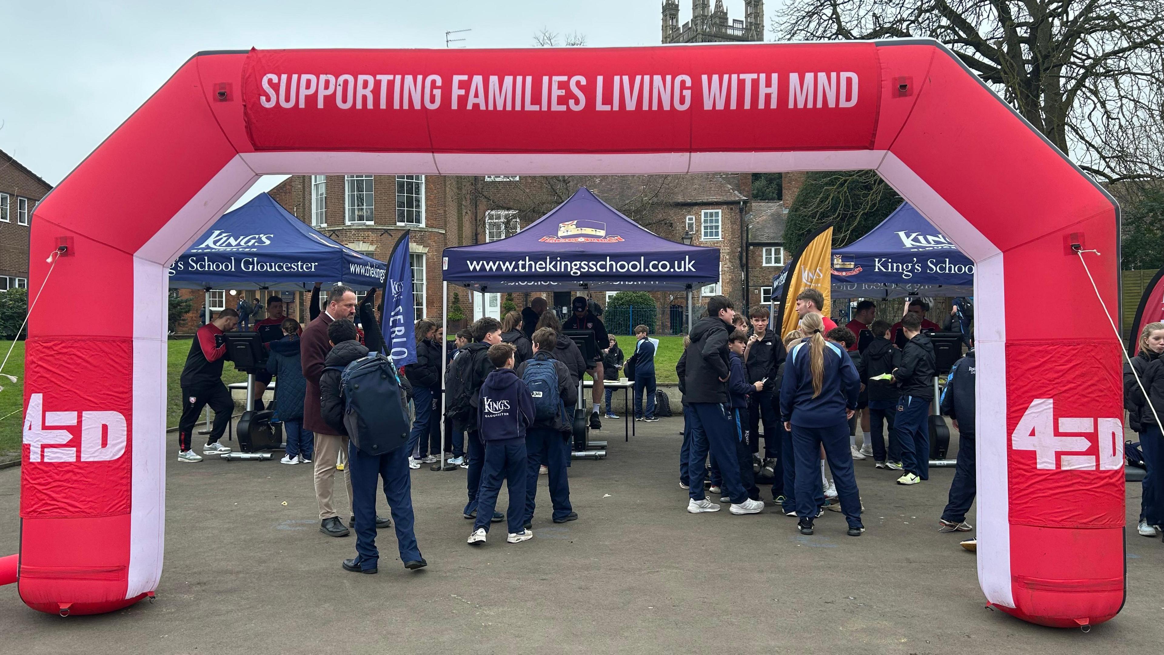 A large red inflatable arch leading towards the three static bikes set up in the school's grounds, underneath individual blue cabanas. There are lots of pupils in PE kits gathered around the bikes, cheering each other on. The inflatable arch says 4Ed on the sides, and 'supporting families living with MND' on the top.