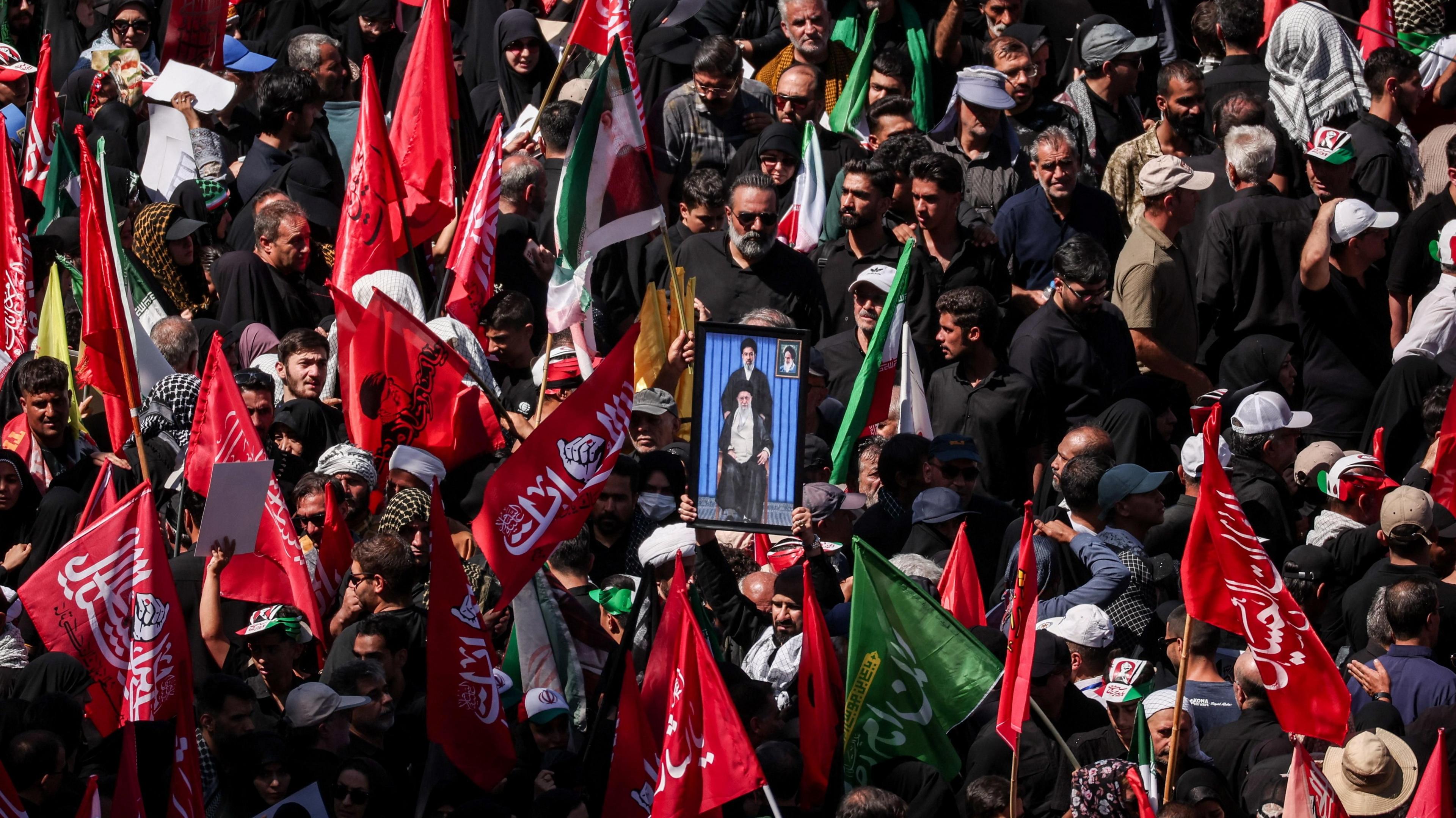 Mourners gather for the burial of Iranian Supreme Leader Ali Khamenei in Mashhad. Many are carrying red flags with Arabic script and in the centre a man holds a picture of a sitting Khamenei, with his son Mojtaba standing above him.