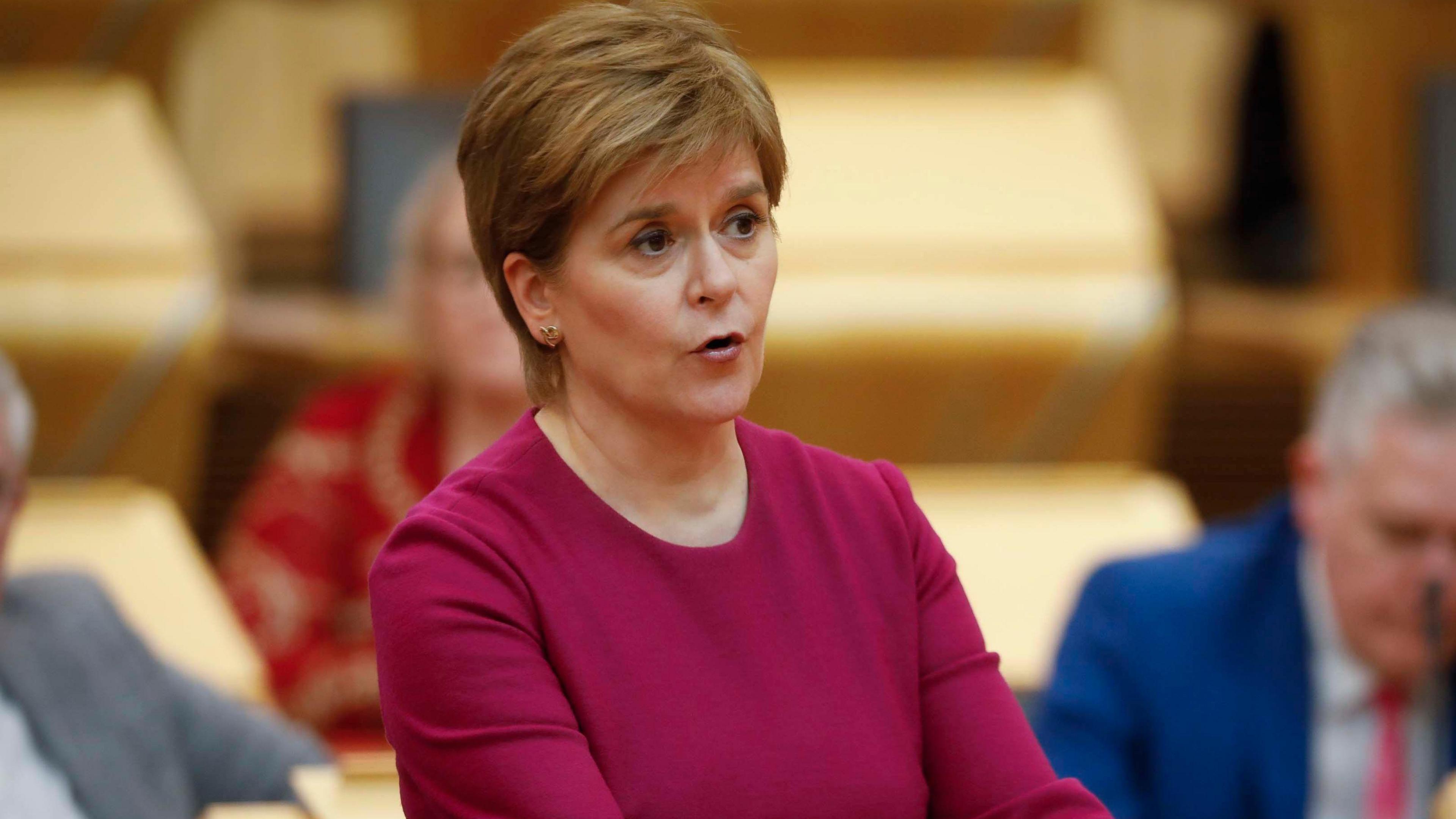 Nicola Sturgeon, who is wearing a maroon dress with a round neck, addresses MSPs in the Scottish parliament debating chamber. She has brown hair, with blond highlights.