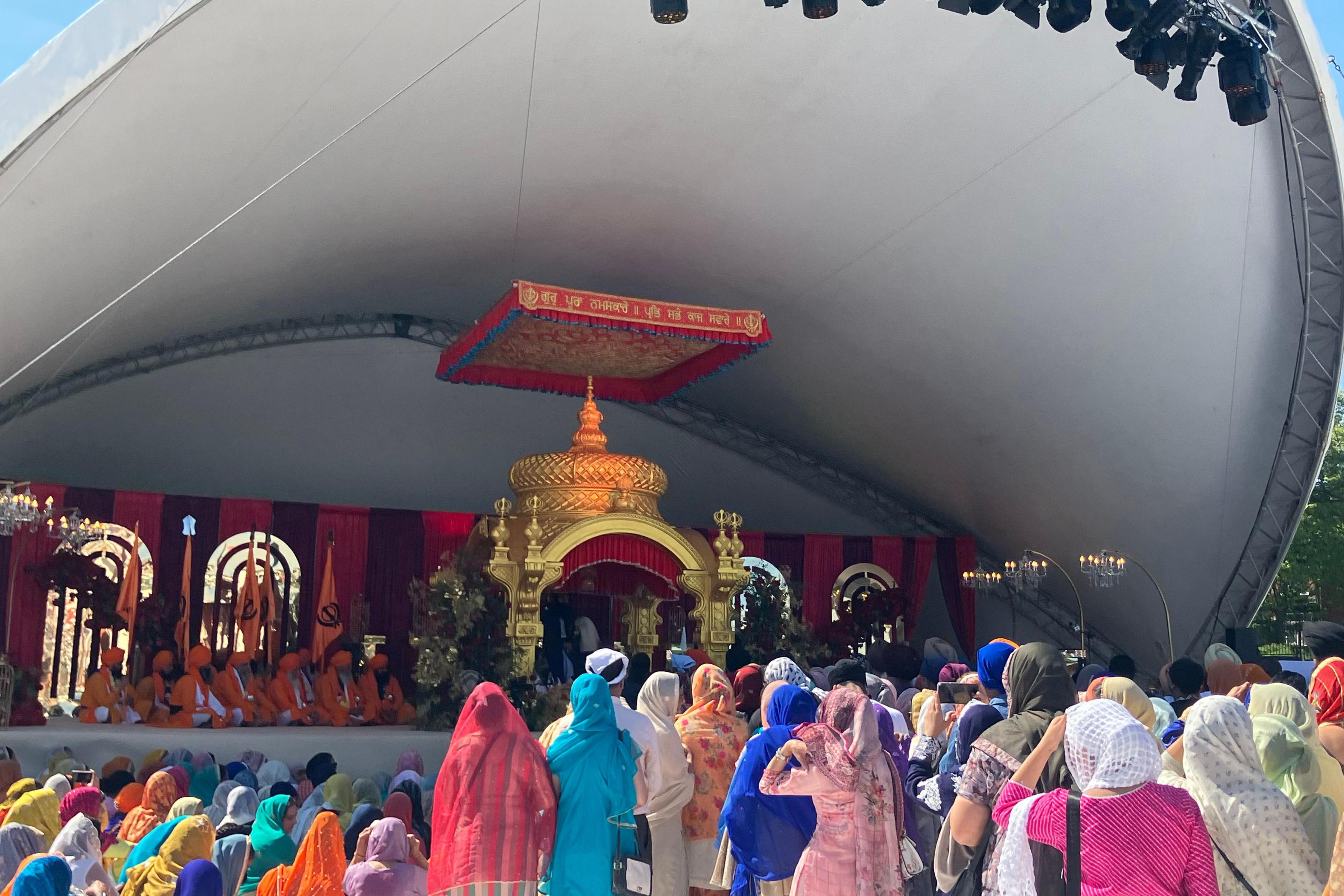 A close up of a large stage with a large gold monument in the centre. To the left of it a group of men in orange traditional South Asian clothing are sitting cross-legged on the stage. In the foreground, a group of people wearing colourful saris are looking towards the stage. 