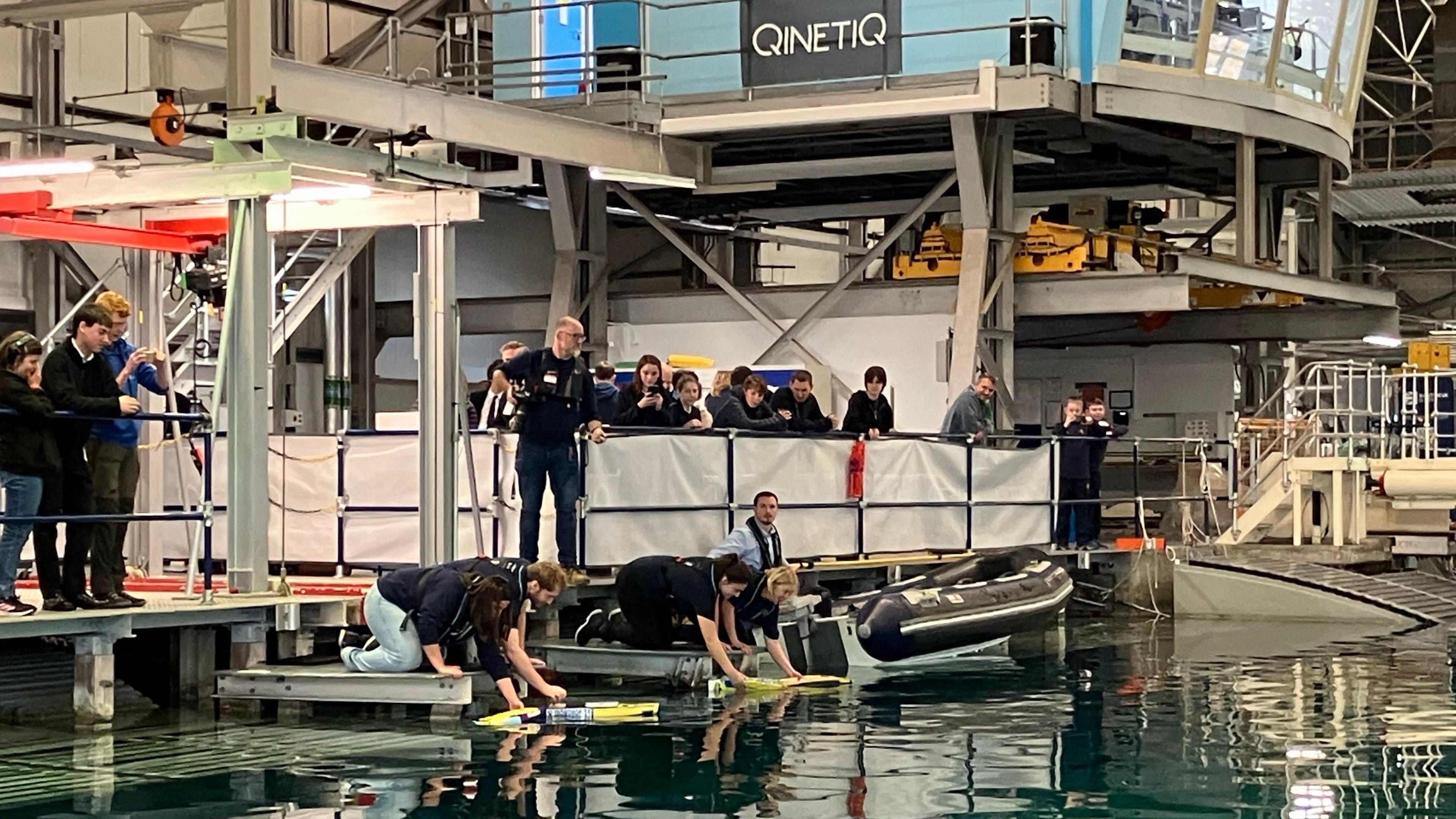 Men and women kneel on platforms above a pool of water at an indoor facility. Their hands are touching small yellow speedboats which are on the surface of the water. There is a crowd of school children watching from a platform above.