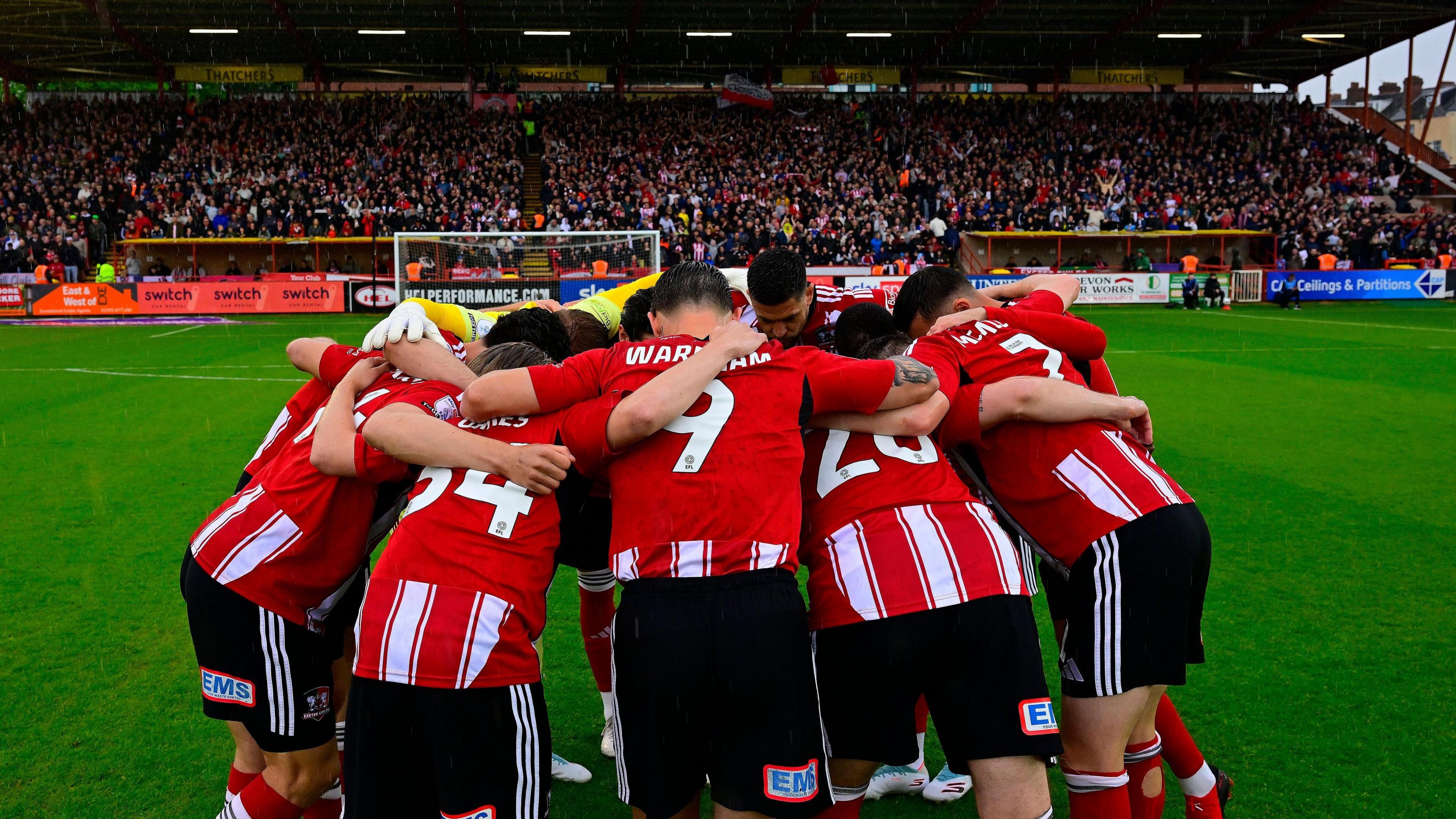 Exeter City players in a huddle in front of the Big Bank