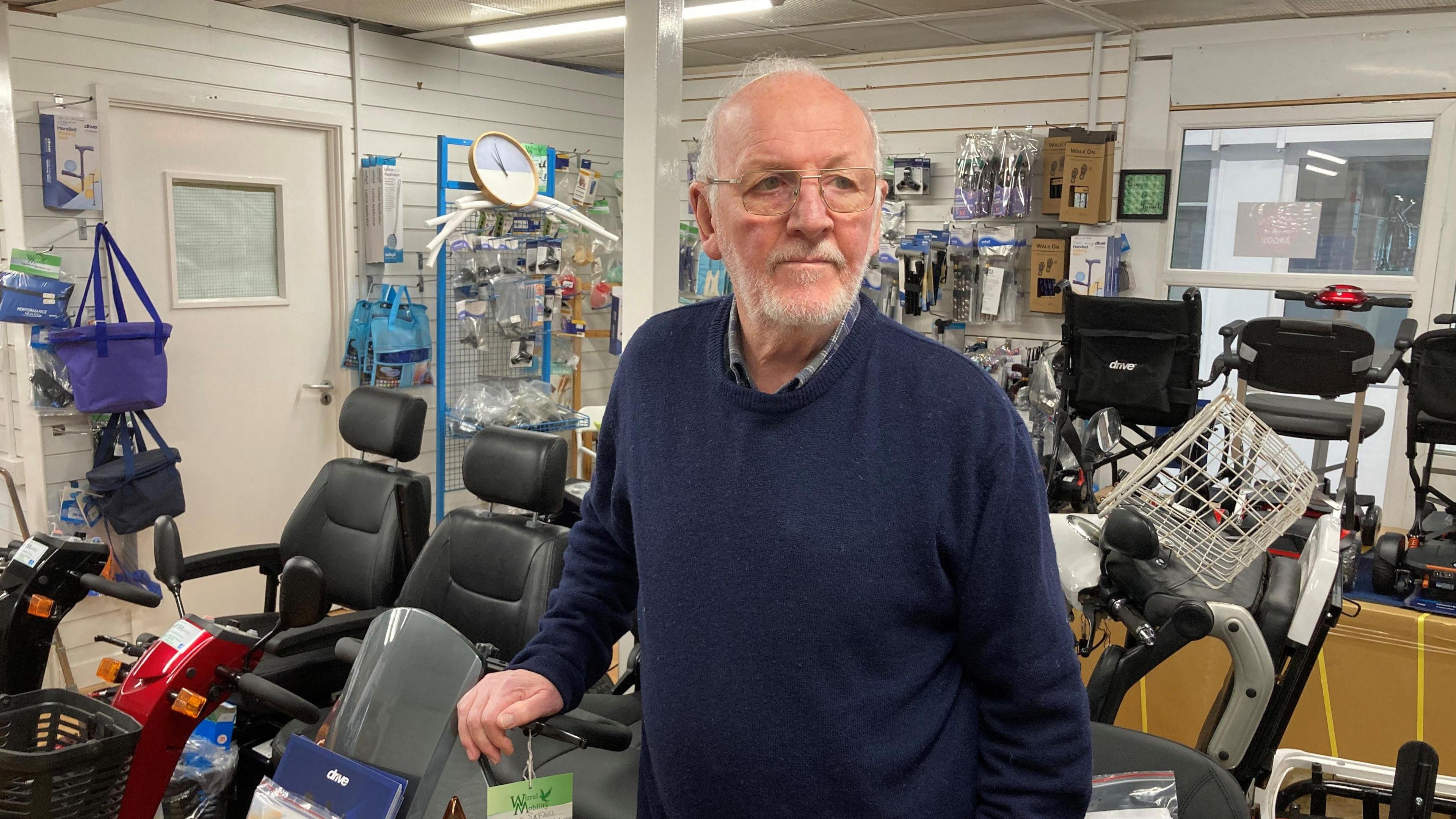 Greg McTigue, who is balding with white hair and wears a blue jumper, stands in a mobility shop surrounded by mobility scooters.