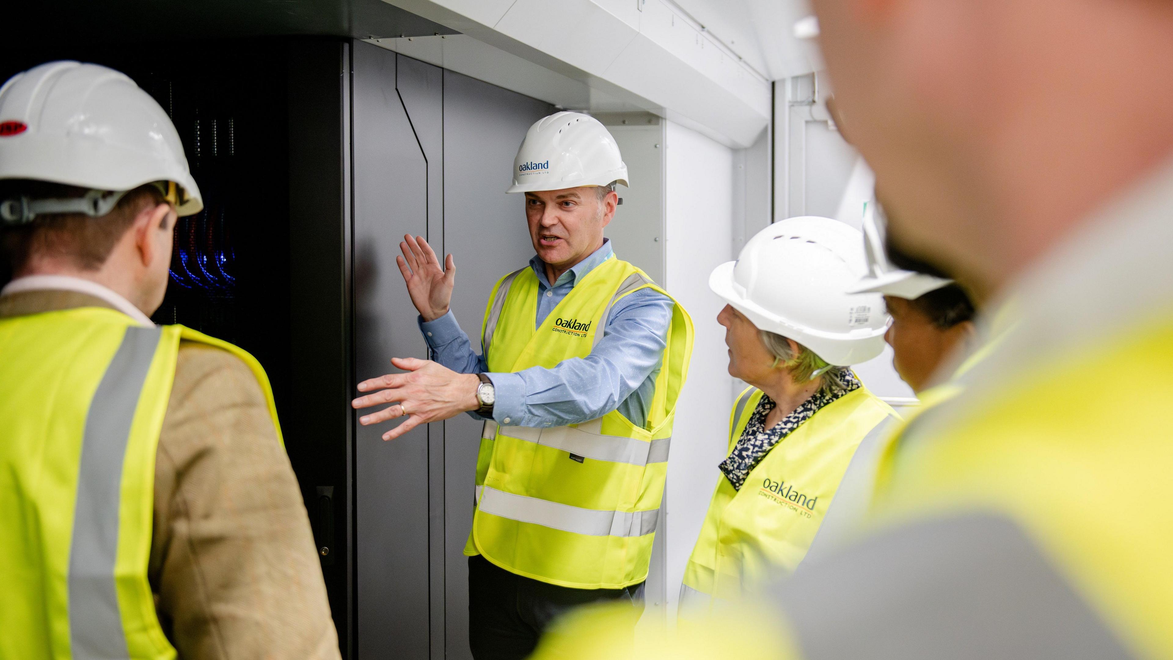 Simon McIntosh-Smith stands next to part of the supercomputer, which is dark grey and white. He is wearing a yellow high-vis jacket and a white hard hat, and appears to be presenting the facility to other people wearing the same safety clothing. 