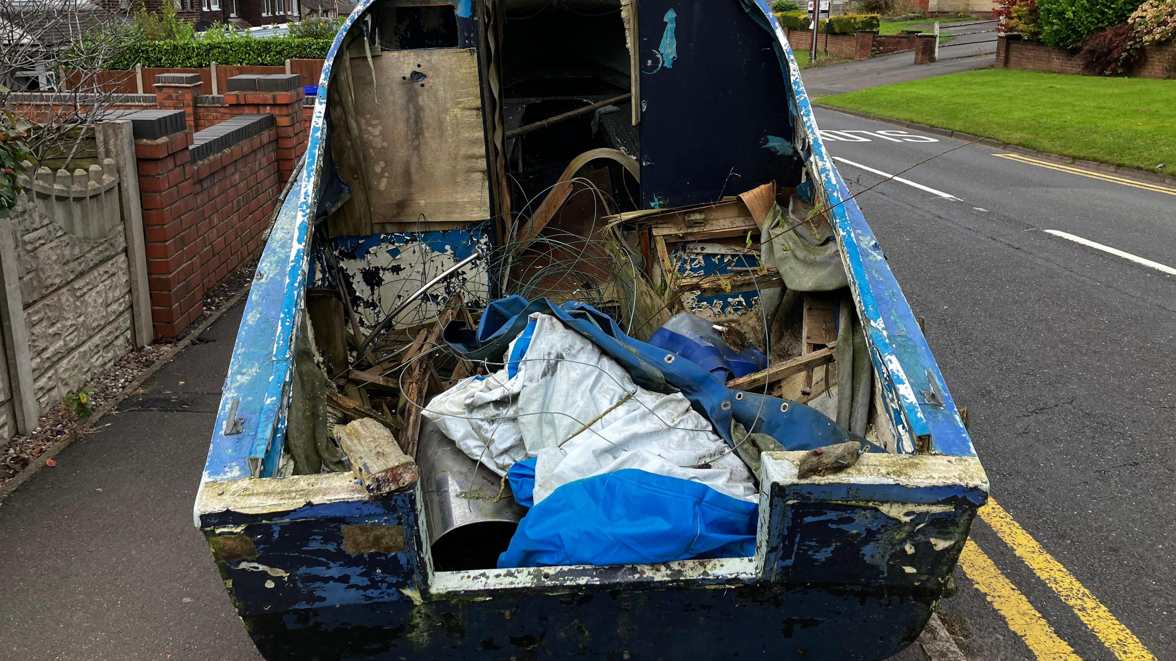 The rear of a blue boat, containing wood, wire, metal and tarpaulin and parked on a pavement