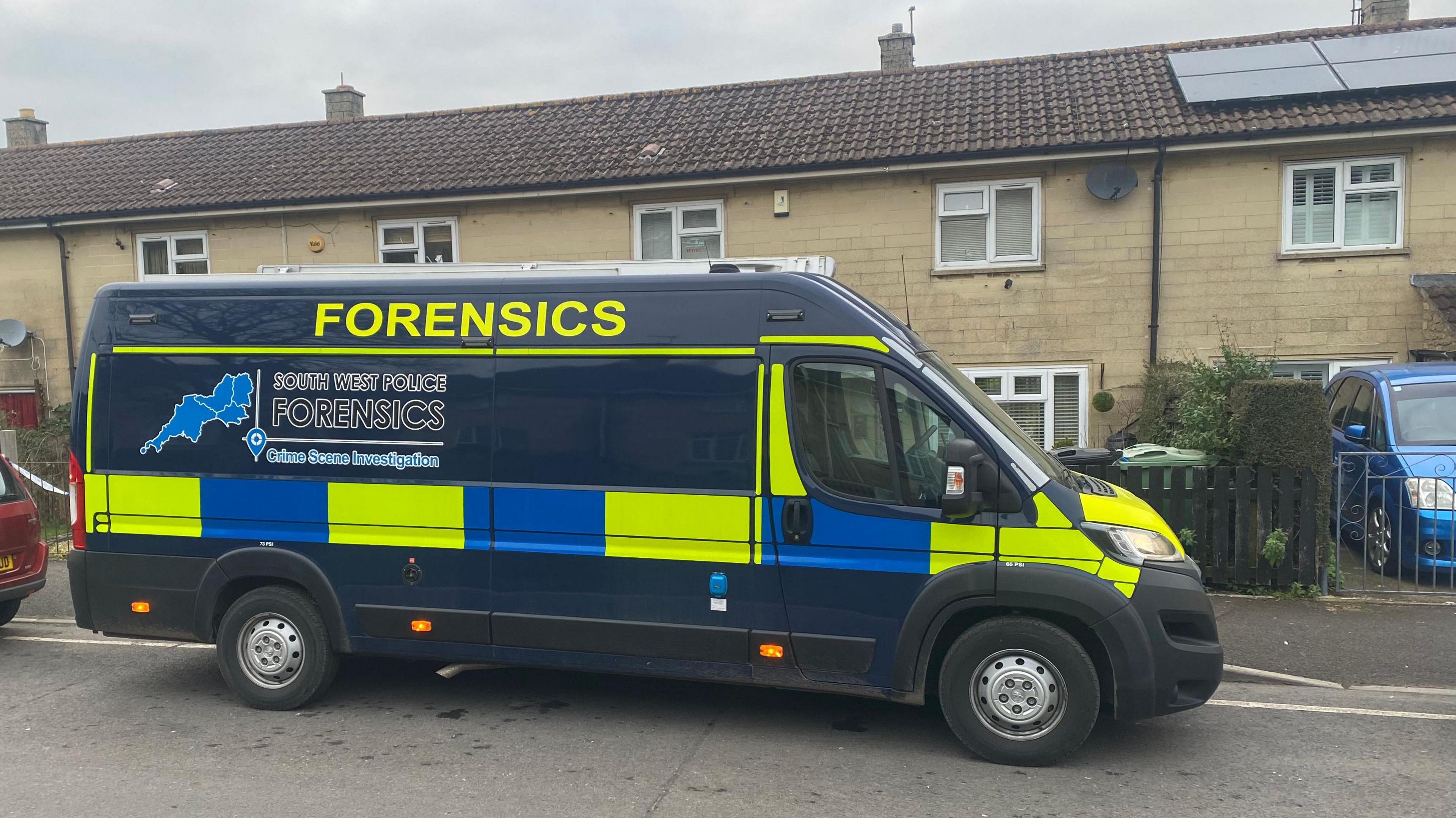 A police forensics van is parked on a road with a row of houses in the background and a red car parked on the left 