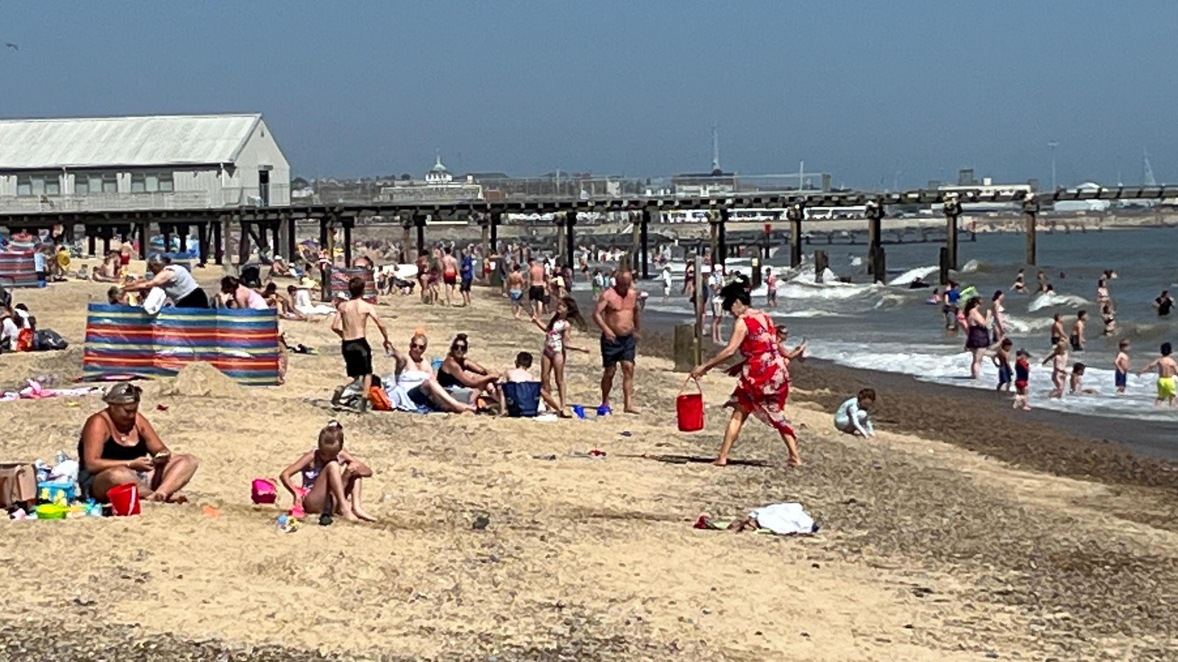 A sandy beach with women, men and children wearing swimming costumes and others paddling or swimming in sea