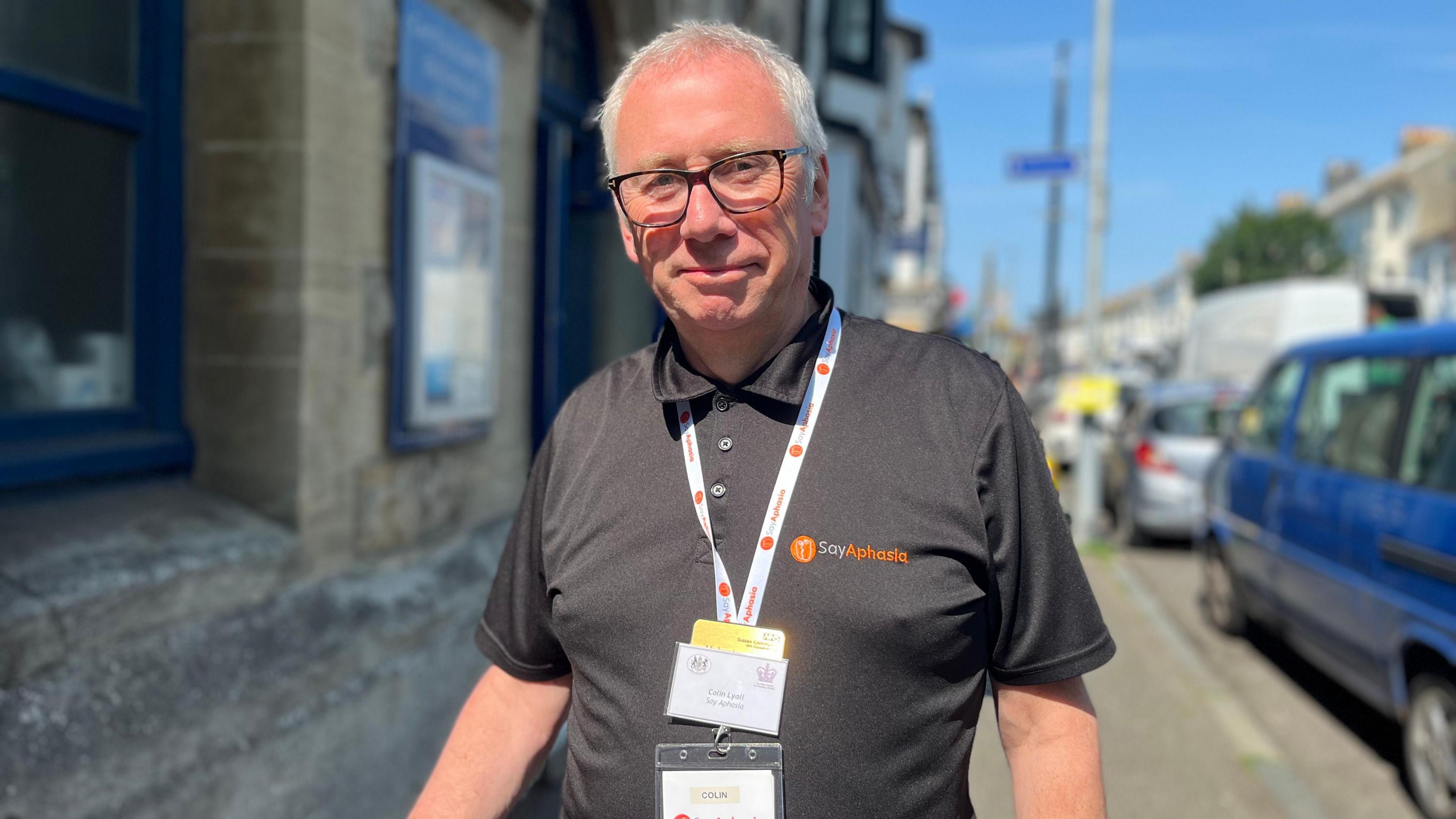 Colin Lyall, a middle-aged man with short receding grey hair and wearing a dark polo shirt, stands on a pavement beside an A-board.
