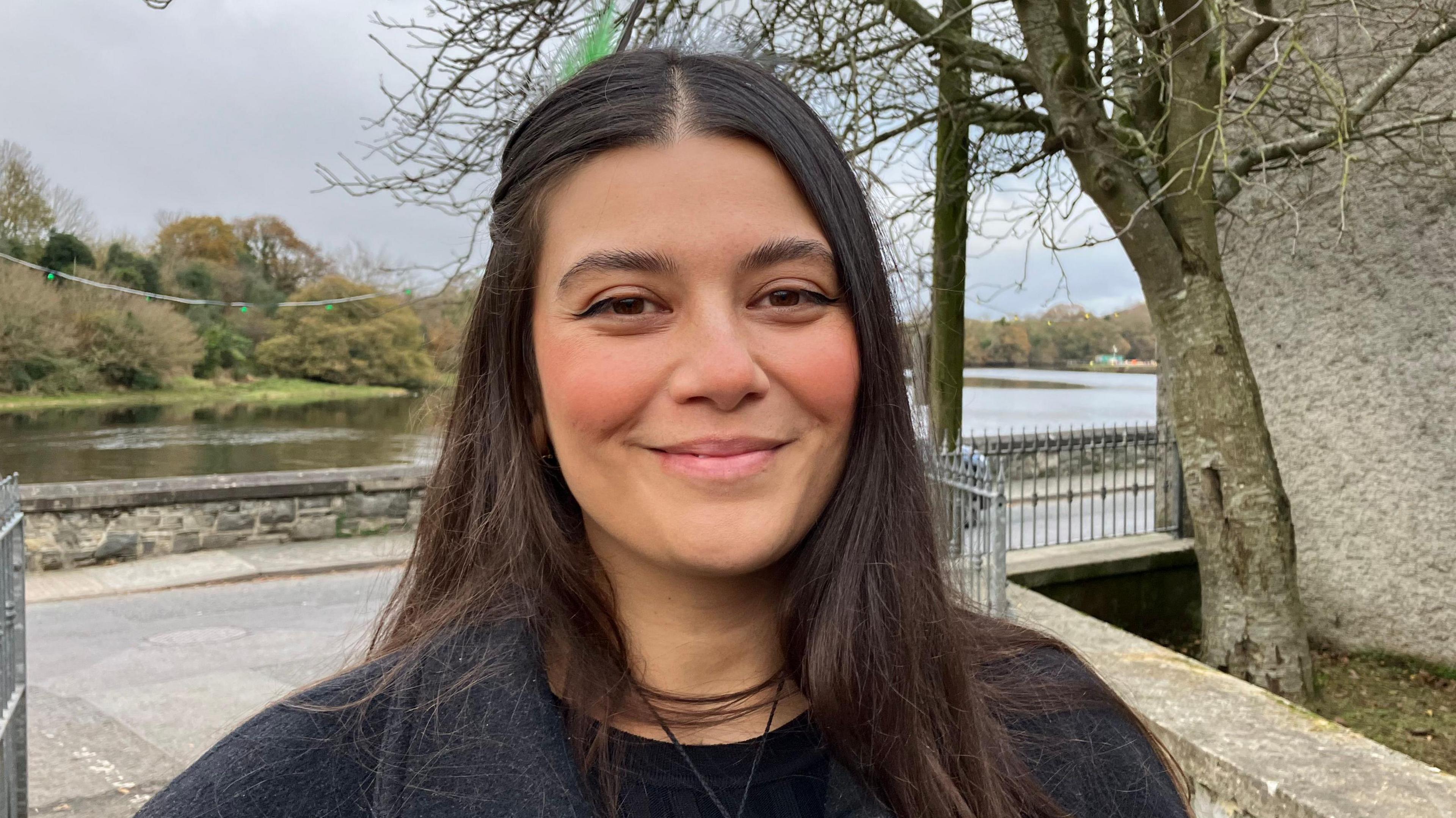 Alyce Ketcher has long dark hair parted in the middle and is smiling to the camera. She is wearing a black coat and black top