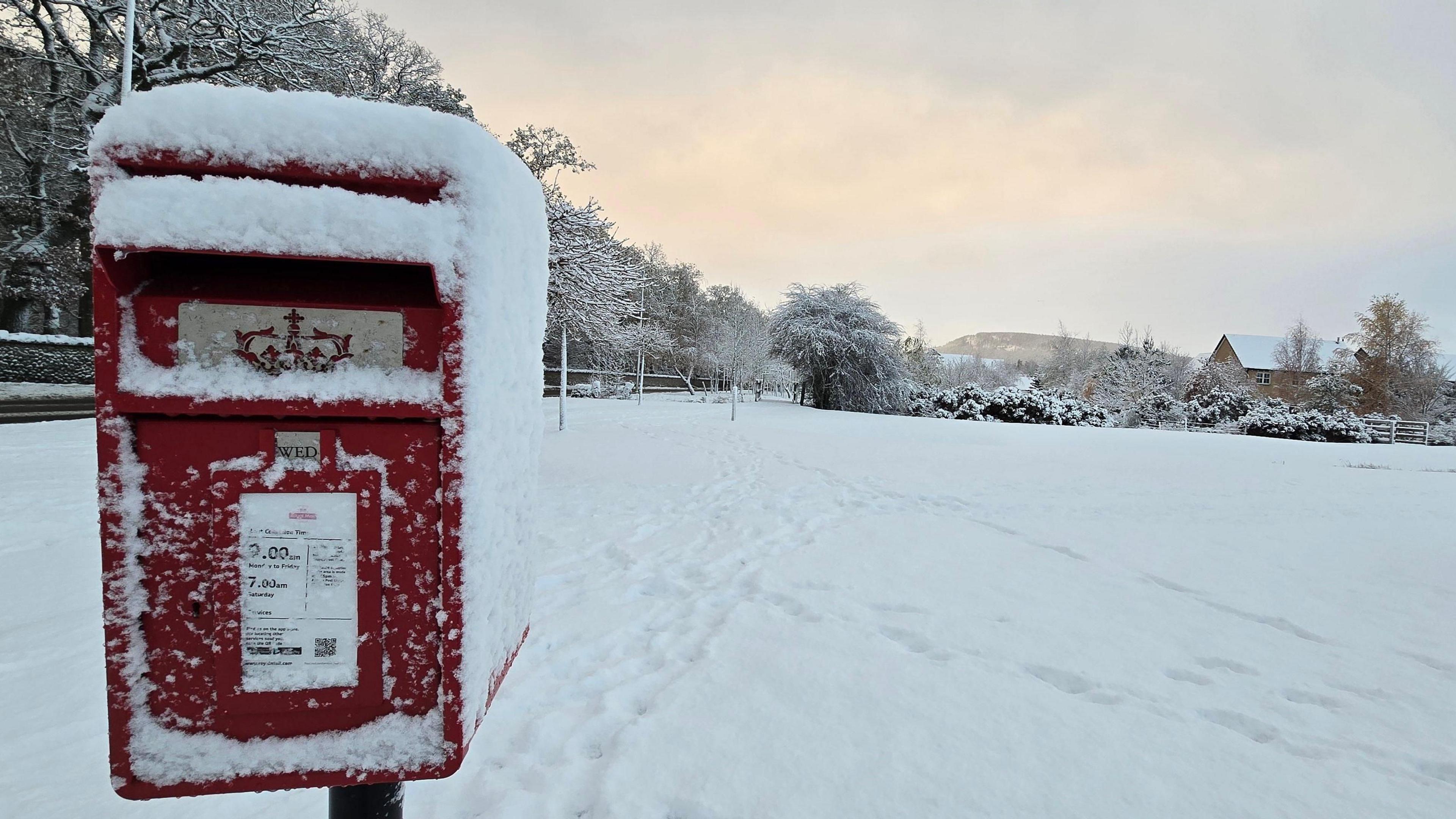 A postbox with snow on its top and sides in the foreground and snow-covered land in the background. 