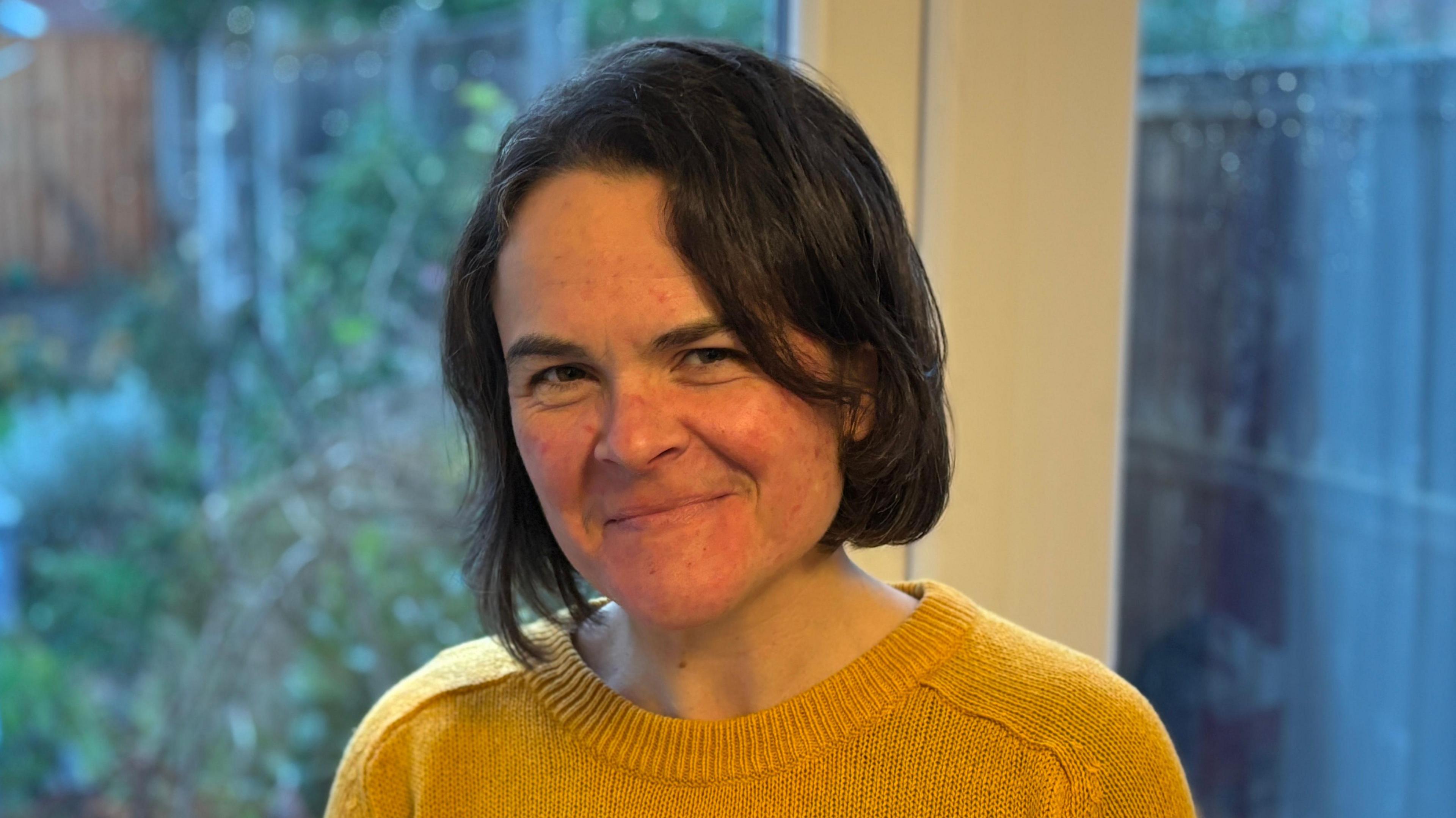 A headshot of a lady with short brown hair, wearing a yellow jumper and standing in front of a white French doors that lead into a garden. She is smiling into the camera 