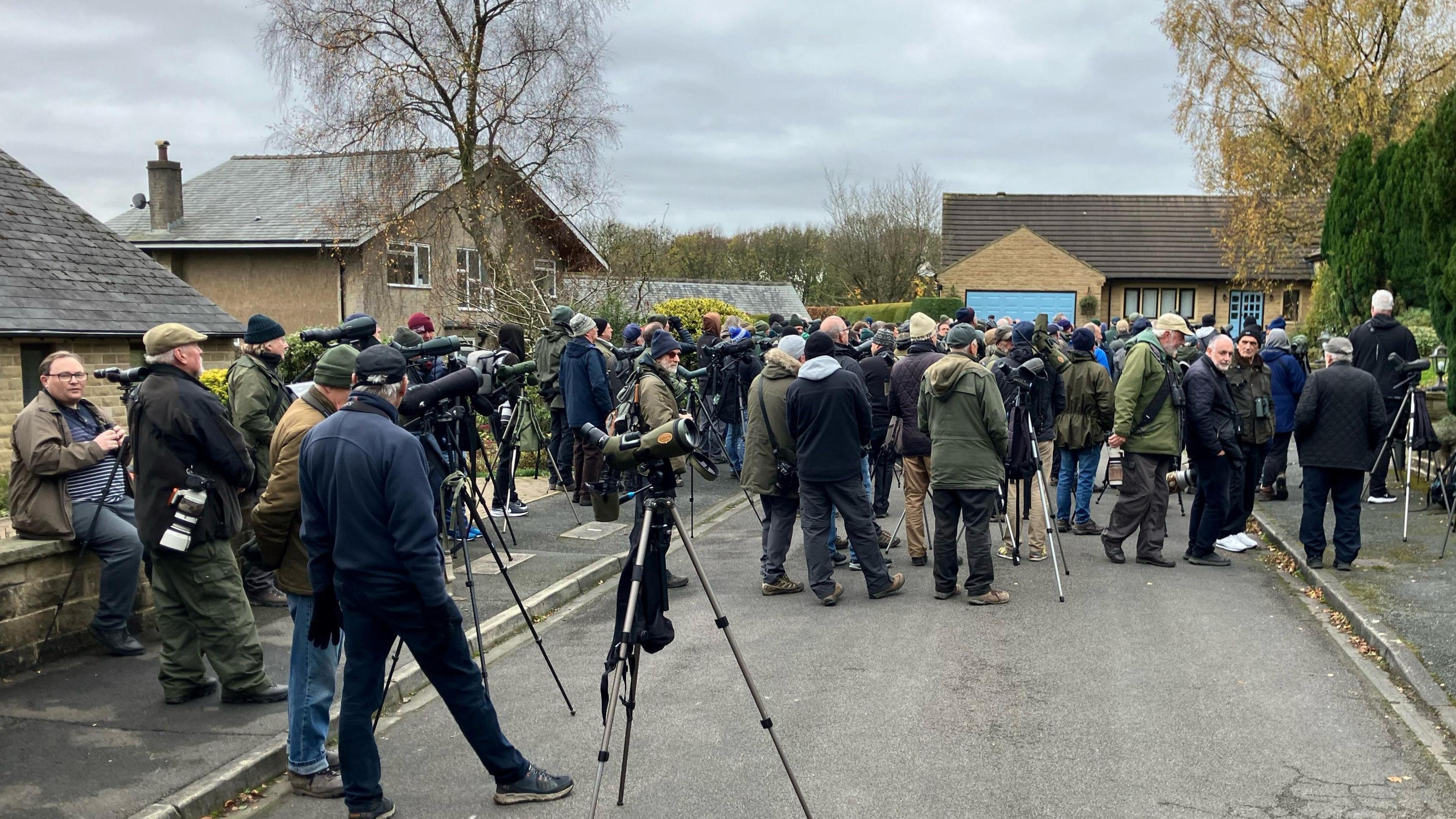 Small residential street with a large crowd of mostly men holding tripods and binoculars dressed in warm coats looking towards one spot