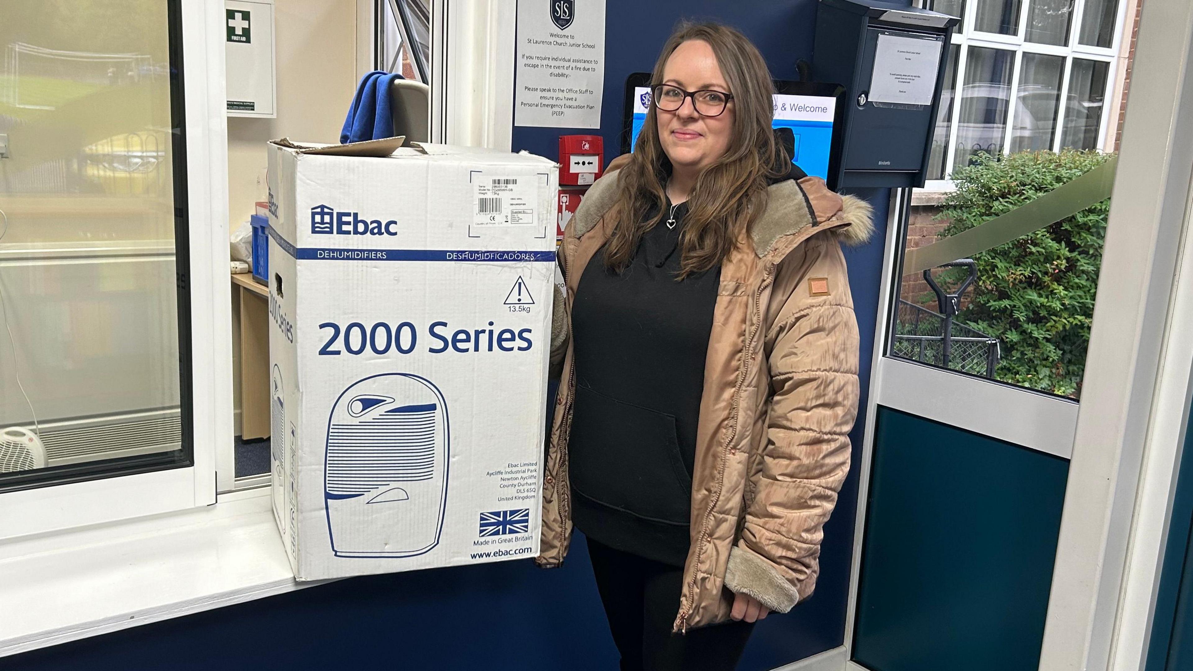 Roxanne Grogan wearing a beige coat holding a box with a dehumidifier inside the school building 