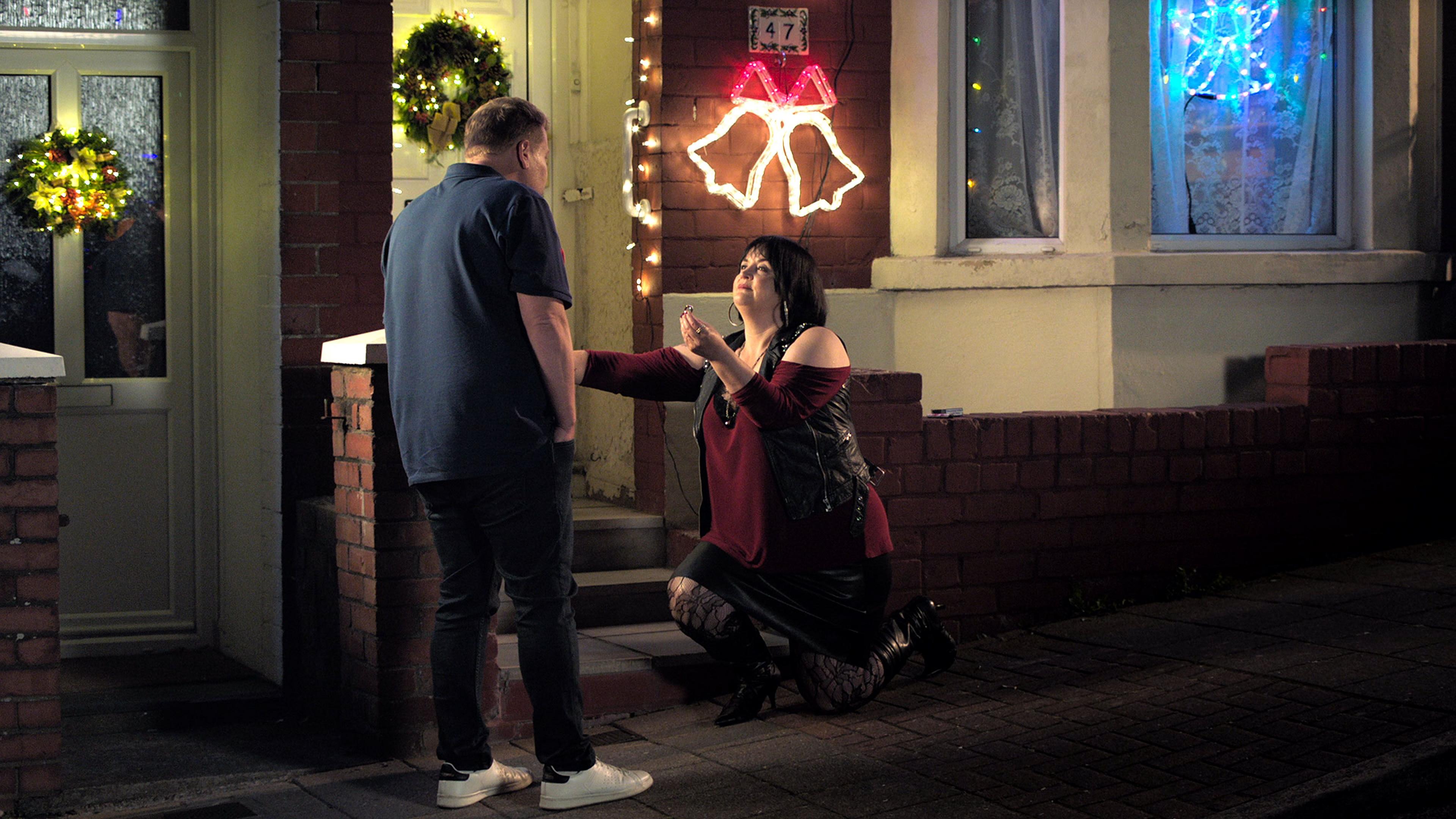 Two people outside a house which painted white and decorated with Christmas lights. The woman, with short black hair and wearing a red top, is knelt down on one knee and holding out a ring. The man, with his back to the camera, is facing her and is blonde, wearing a navu polo shirt and jeans.