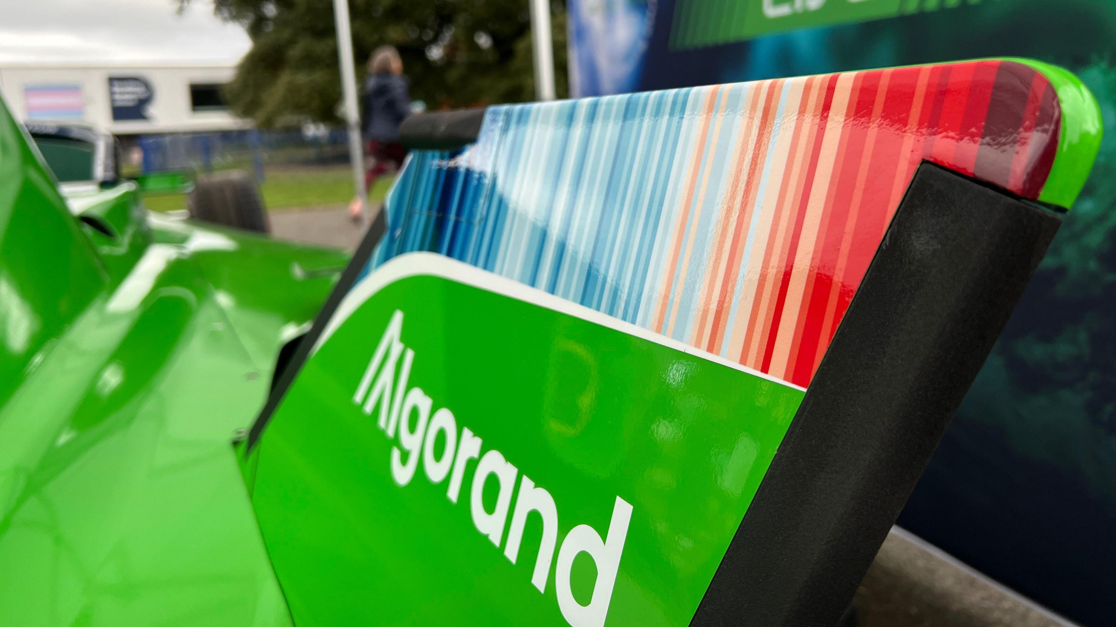The image shows a close up of the climate stripes featured on the rear wing of the race car. It goes from dark blue on the left side to pale blue in the middle and then red and dark red on the right side. The car is a bright green.