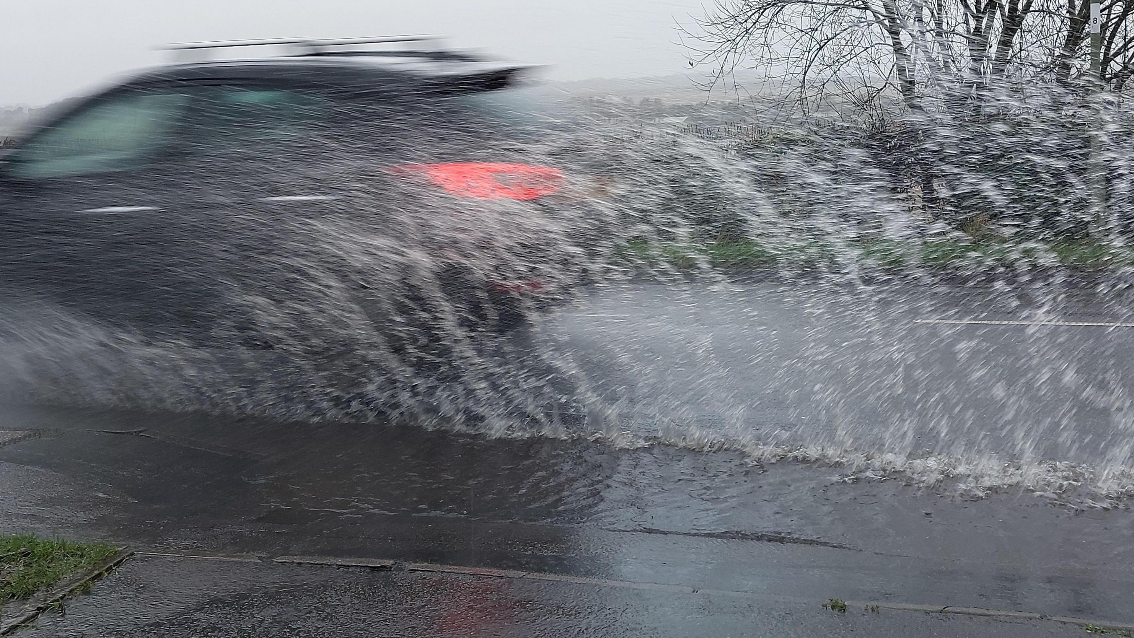 A car drives through a puddle, leaving a spray of water in its wake.