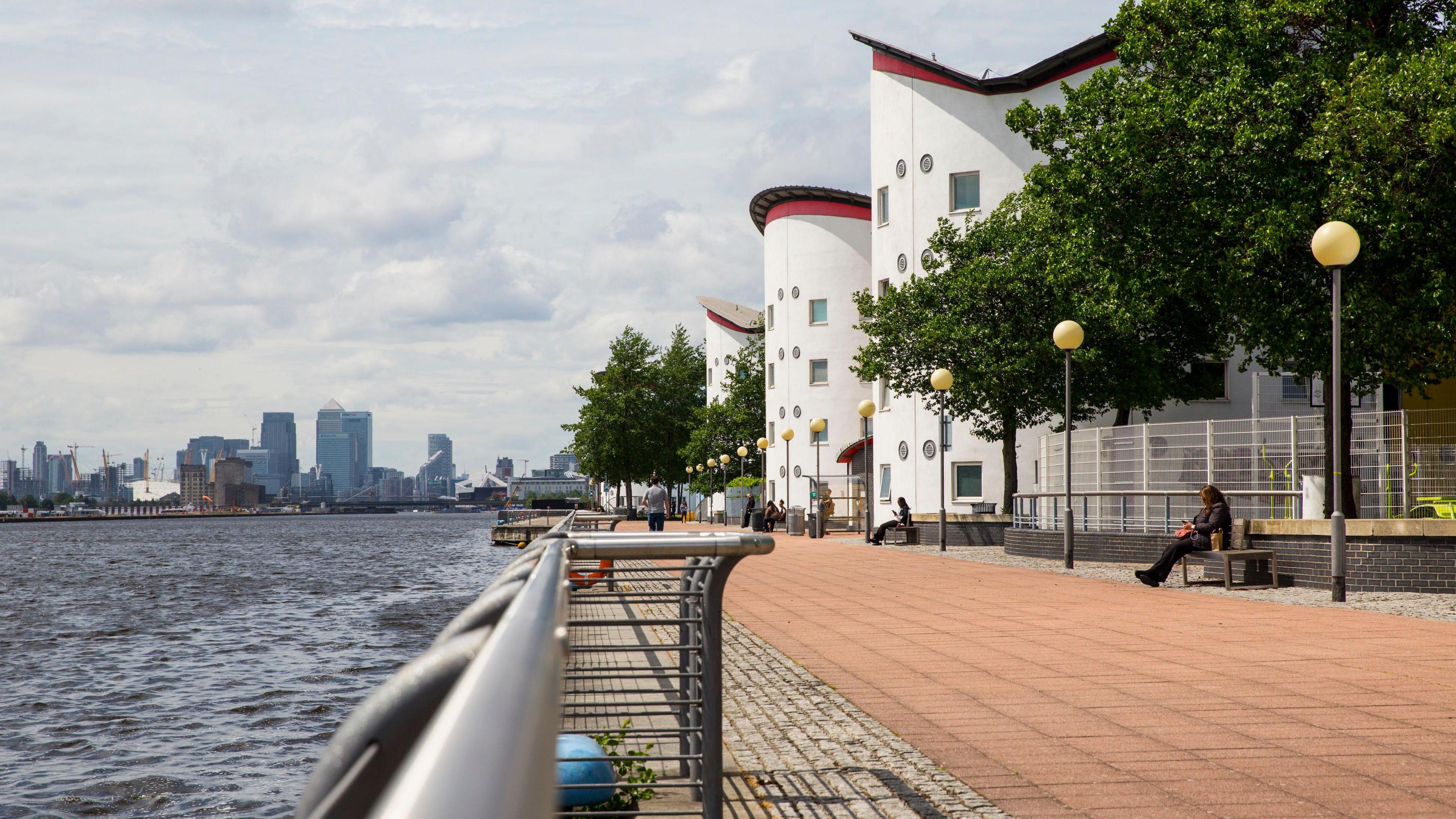 The River Thames with Canary Wharf in the distance and a path along the river past trees and white, cylindrical buildings that is student accommodation at the Docklands campus of the University of East London