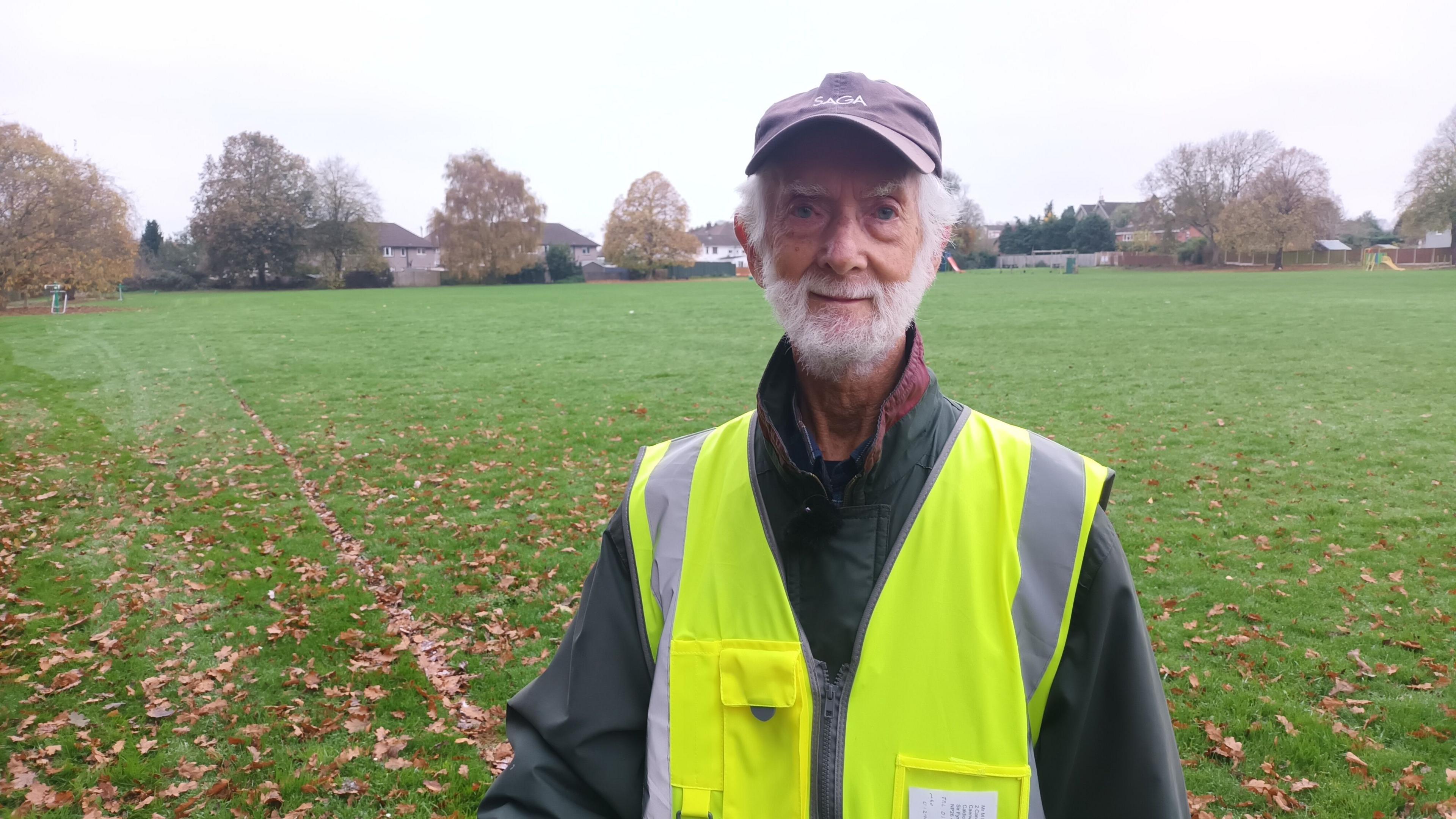 Michael Barnfather wearing a hat and a yellow high-vis jacket, smiles at the camera as he stands in a field 