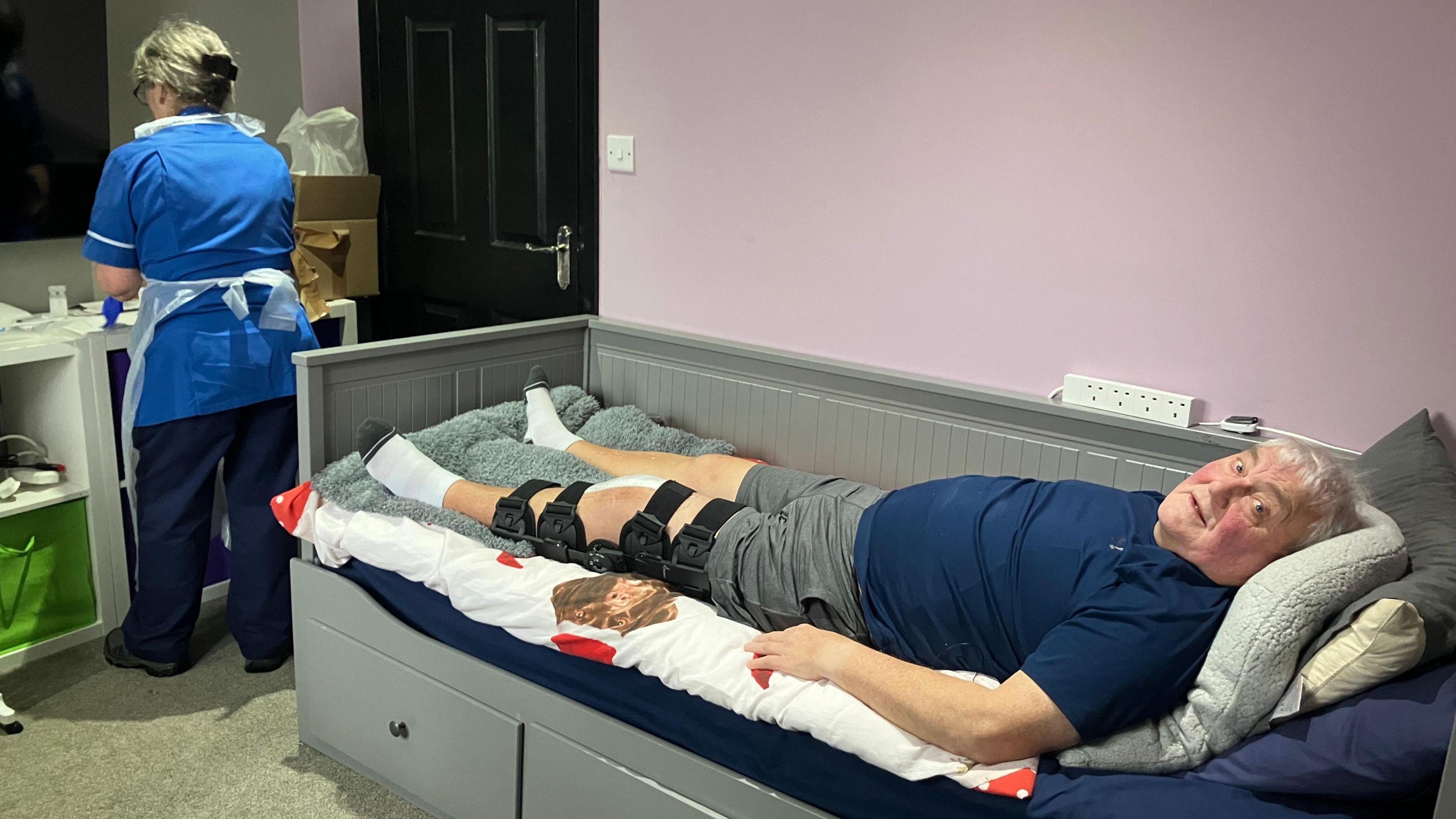 Wayne Duffey, with a brace on his knee, lays down on a single bed. A nurse in a blue uniform attends. 