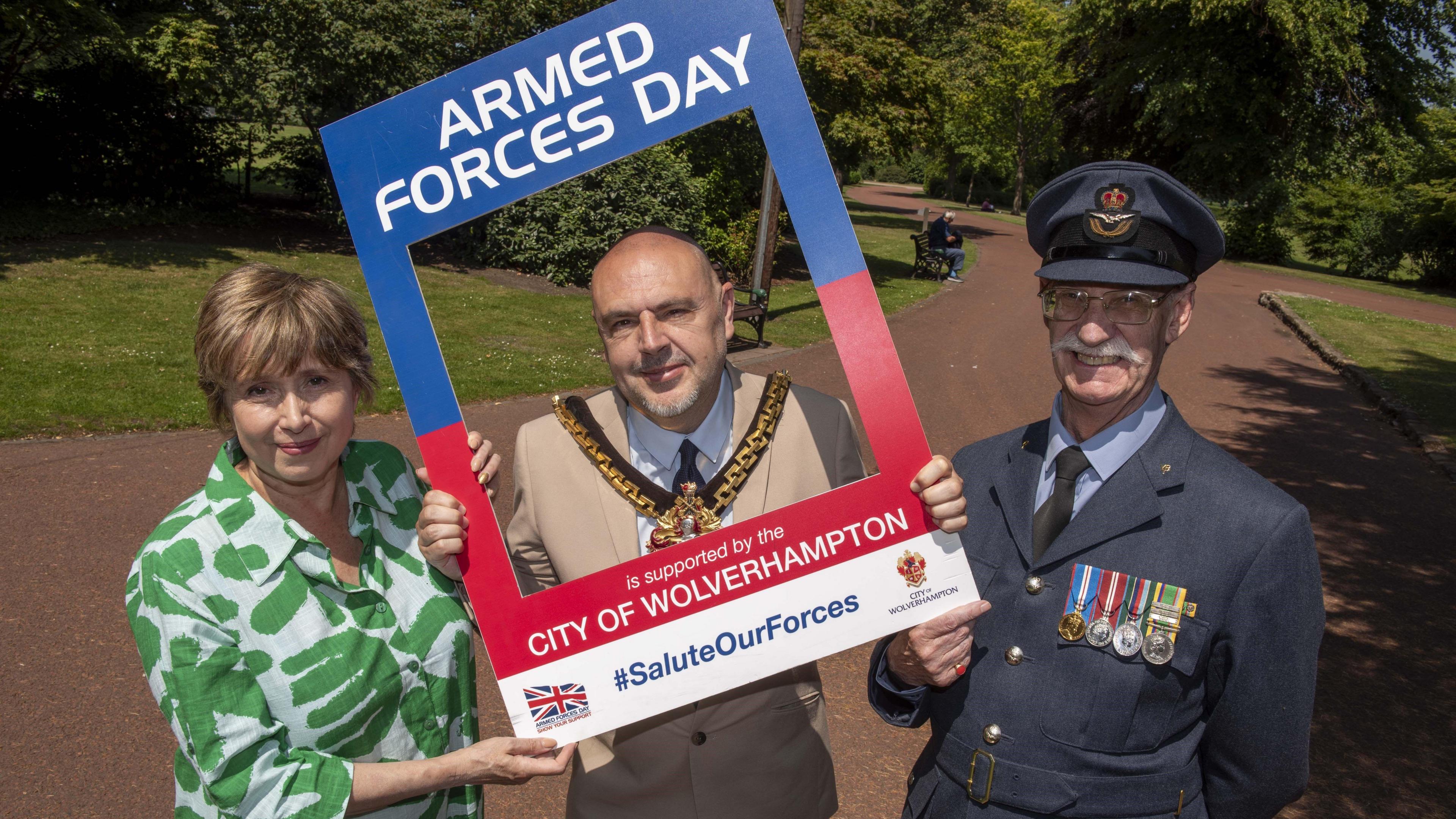 A woman and two men are standing in a park which has green grass and a brown path. They are holding a carboard with a square cut out. One of the men is standing in the cut out. It reads "armed forces day is supported by the city of Wolverhampton." The man in the middle is the mayor and is dressed in a beige suit with a black and gold mayoral chain. The man next to him is wearing a navy coloured armed forces uniform, with four badges on his breast.