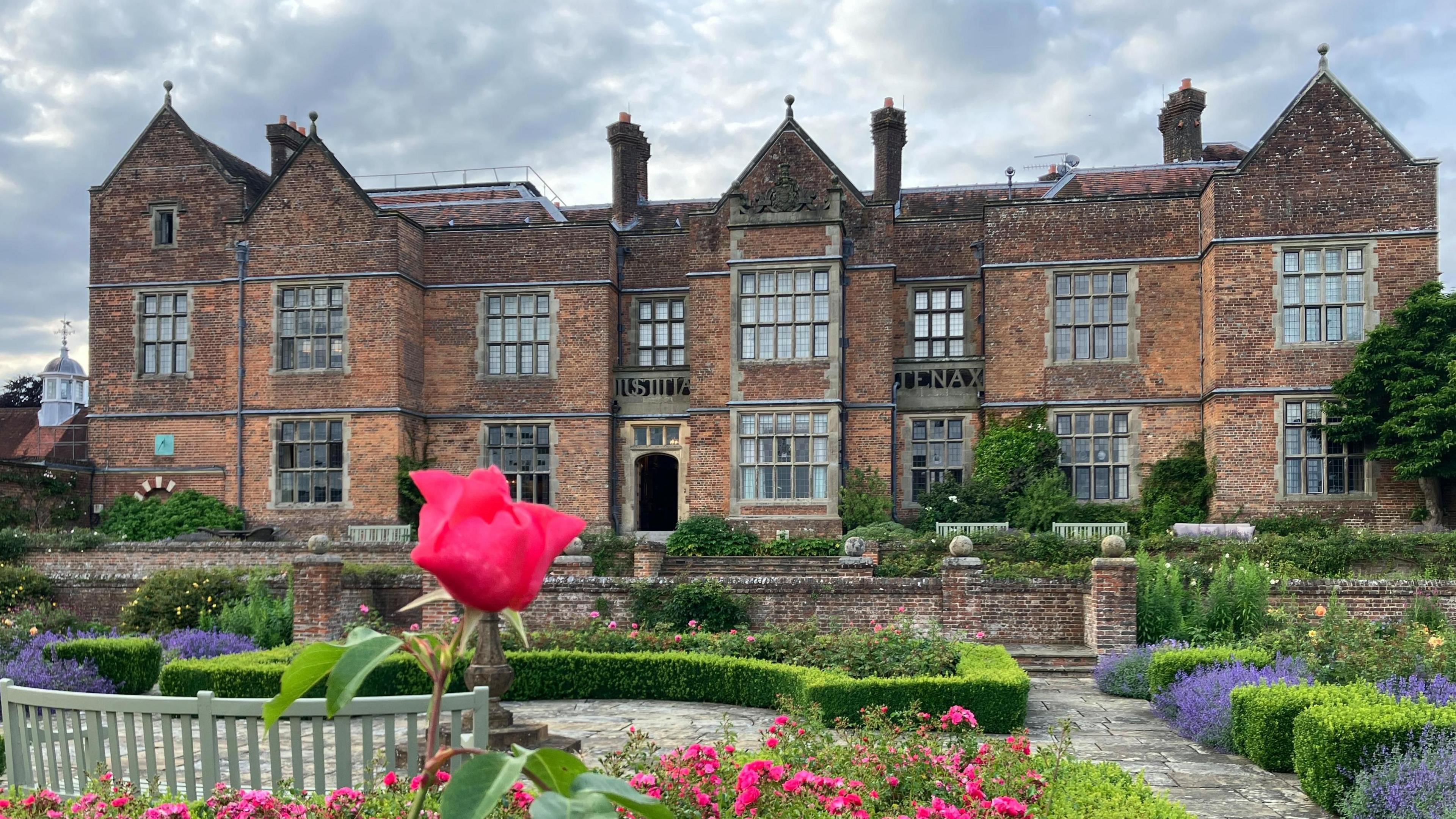 Chequers a large 16th century manor house with a garden to its front. The house has numerous brightly-coloured flowers in its garden and a bench facing the entrance of the house. 