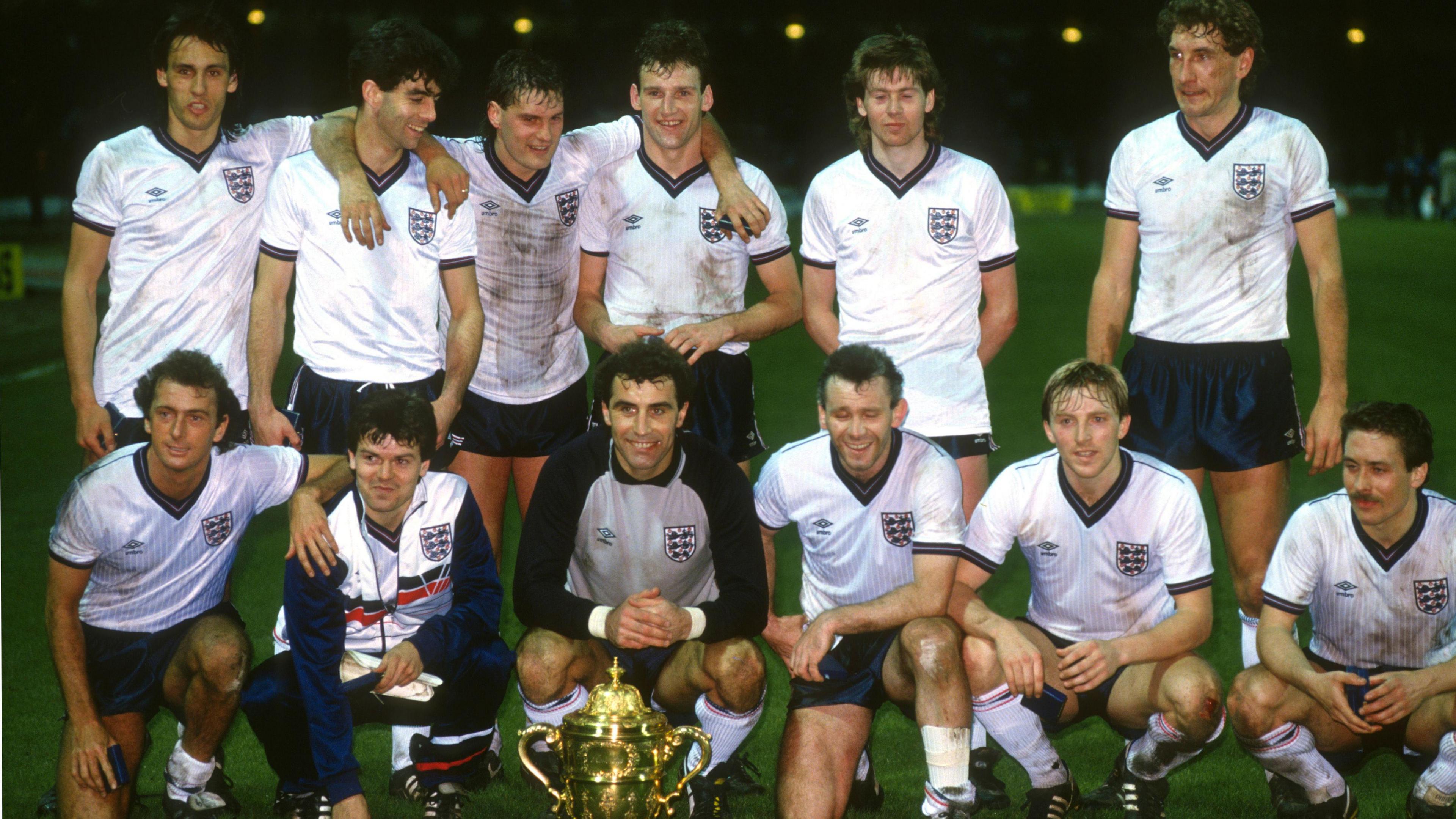 An England team photo, with 13 men dressed in white shirts and blue shorts lined up in two rows on a green pitch following a match. Players in the back row have their arms around one another, while those in the front row are kneeling behind a large gold trophy.