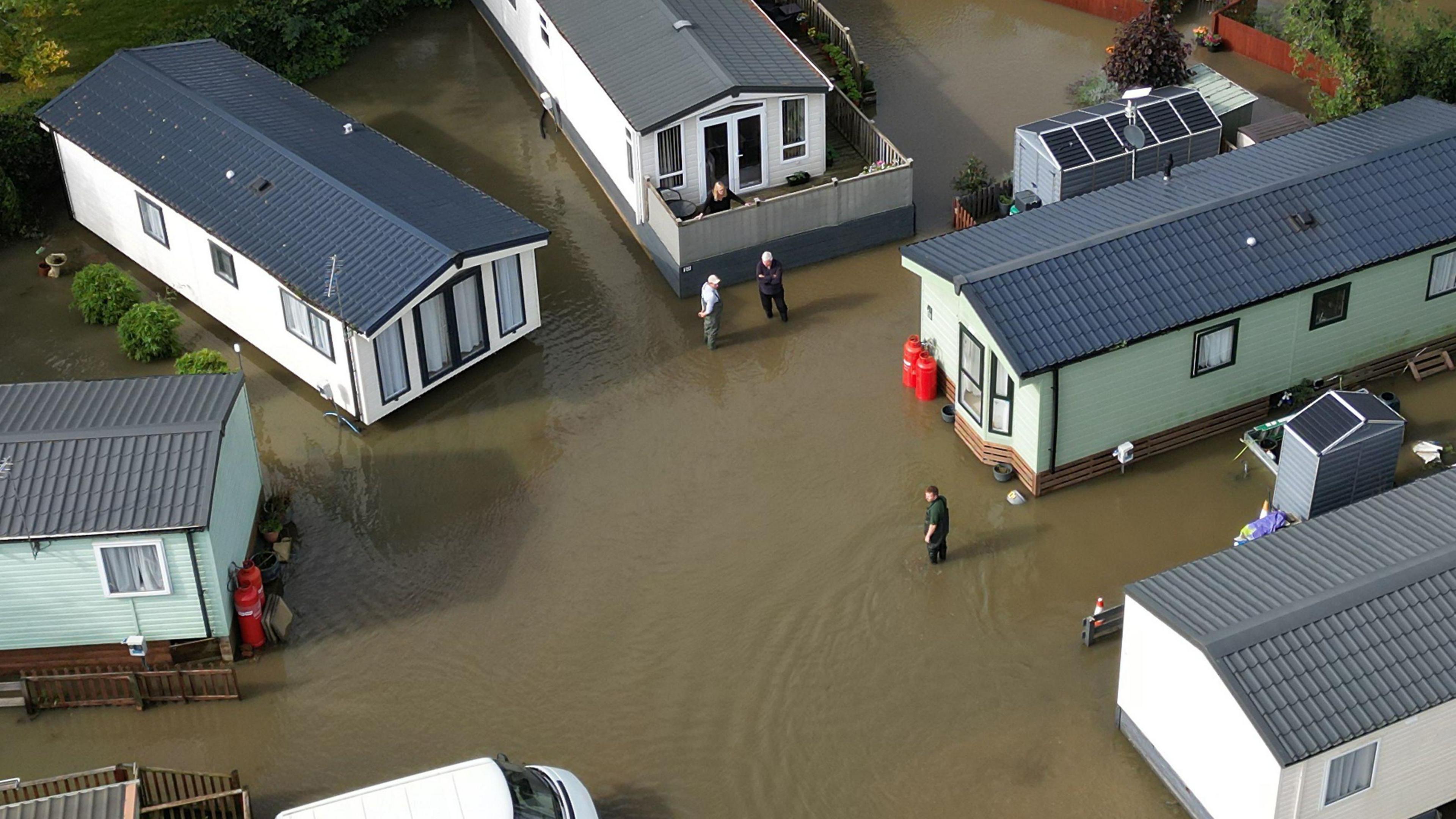 Another drone image of flooding in Cogenhoe, this time showing five static caravans which are either white or light green in colour. A few people are standing in brown water that, in terms of depth, is just under their knees. The water is totally surrounding each caravan.