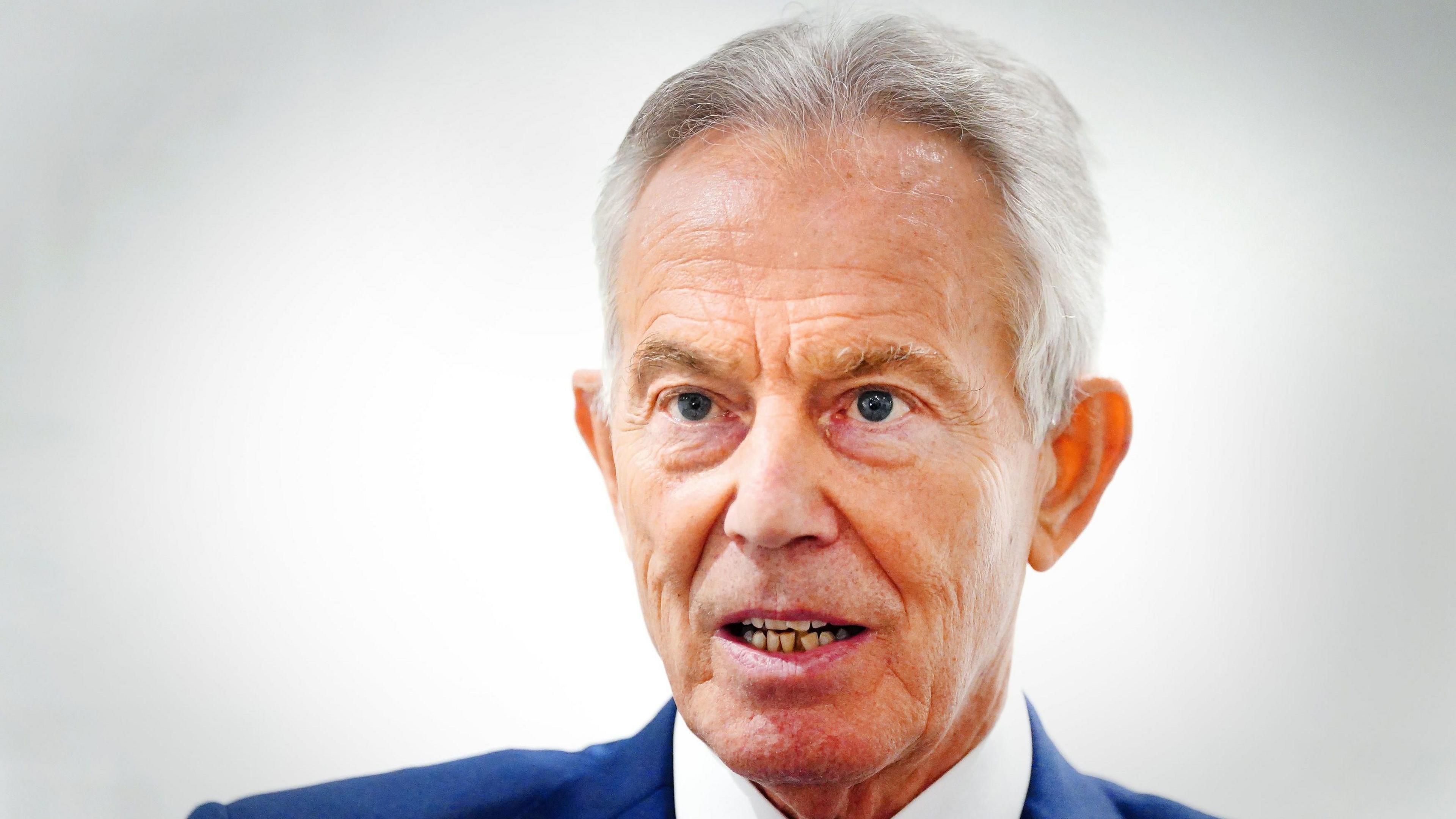 Tony Blair has white hair and is wearing a dark blue suit jacket with a white shirt underneath. He is speaking and looking into the distance
