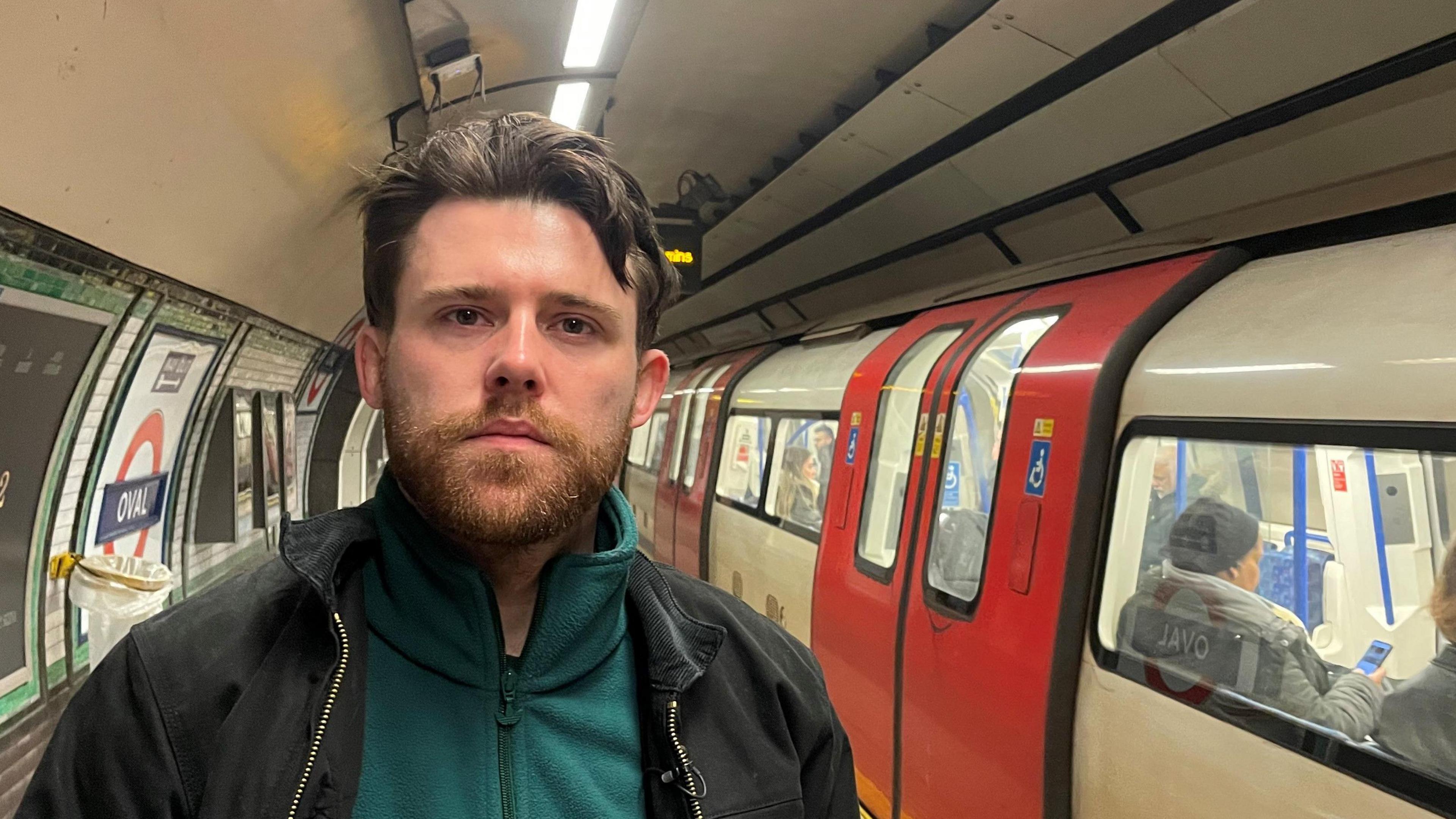 Niall McNamee, standing on a London underground platform while a tube passes on the left-hand side. Niall has dark, short hair, and a beard. He is wearing a black jacket, with a green fleece underneath.