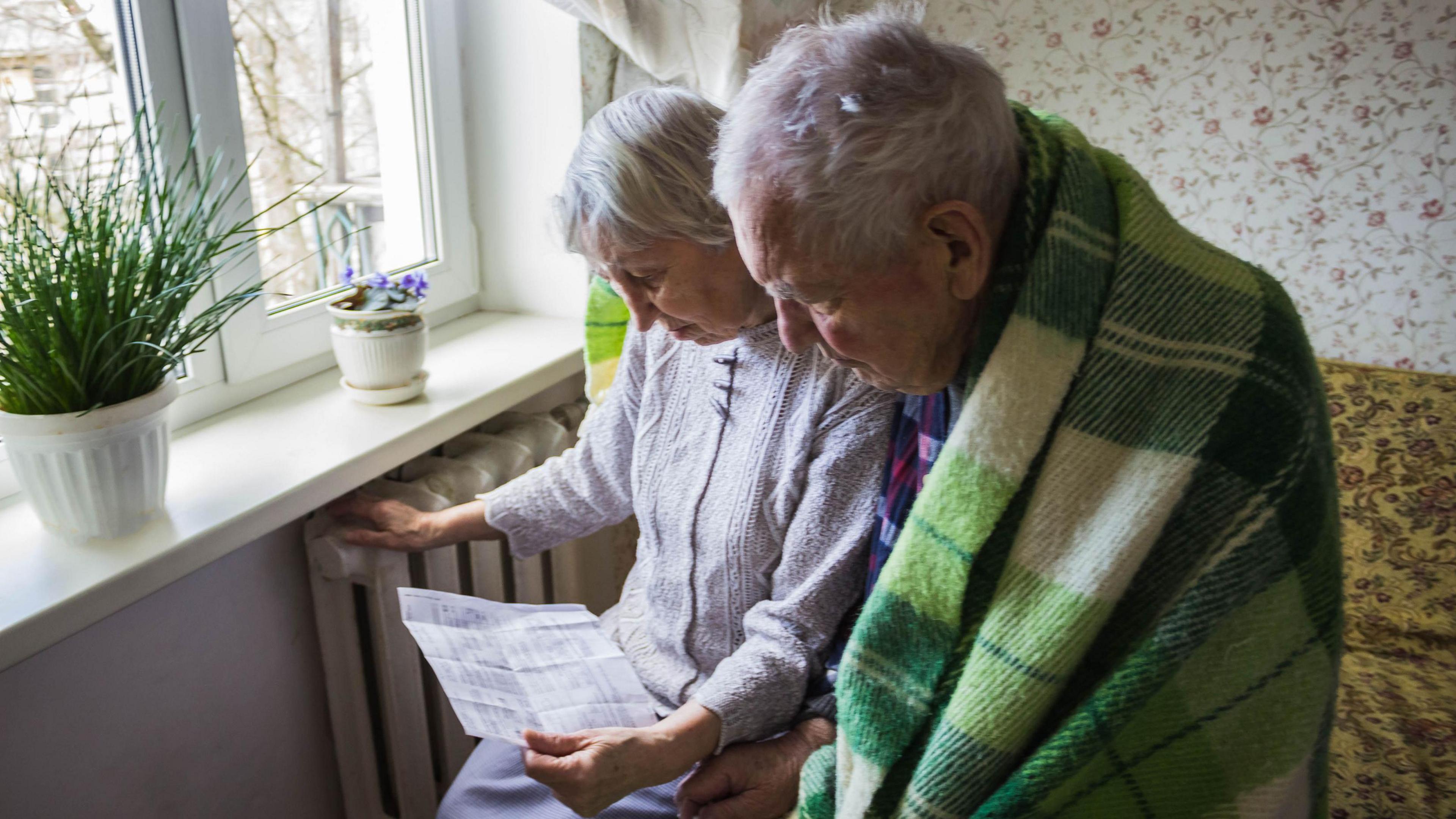 Two pensioners sat near a radiator looking at heating bills. One of them has a blanket around him. 