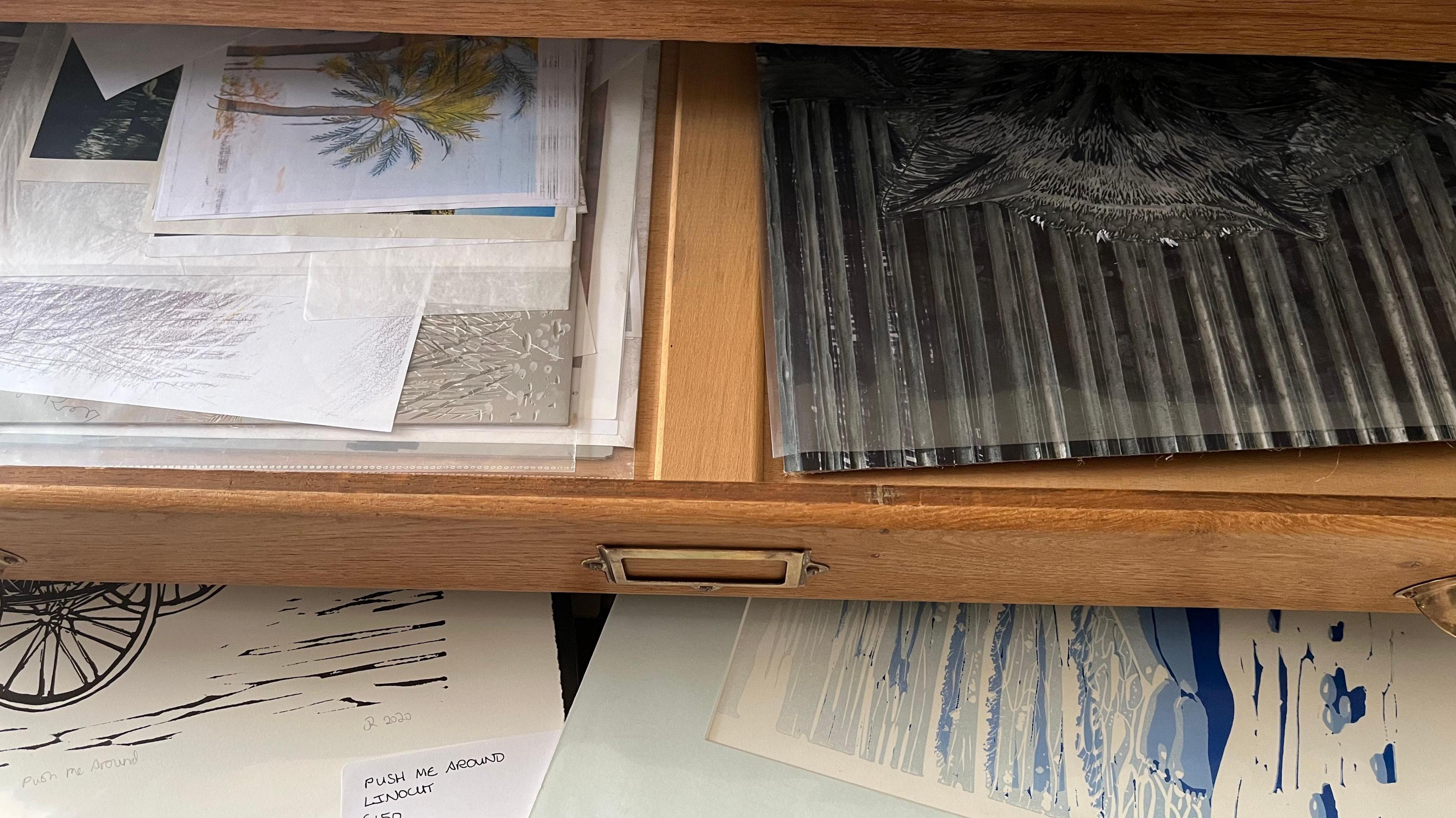 An open pine wood drawer - inside are various lino-cut illustrations in black and blue ink.