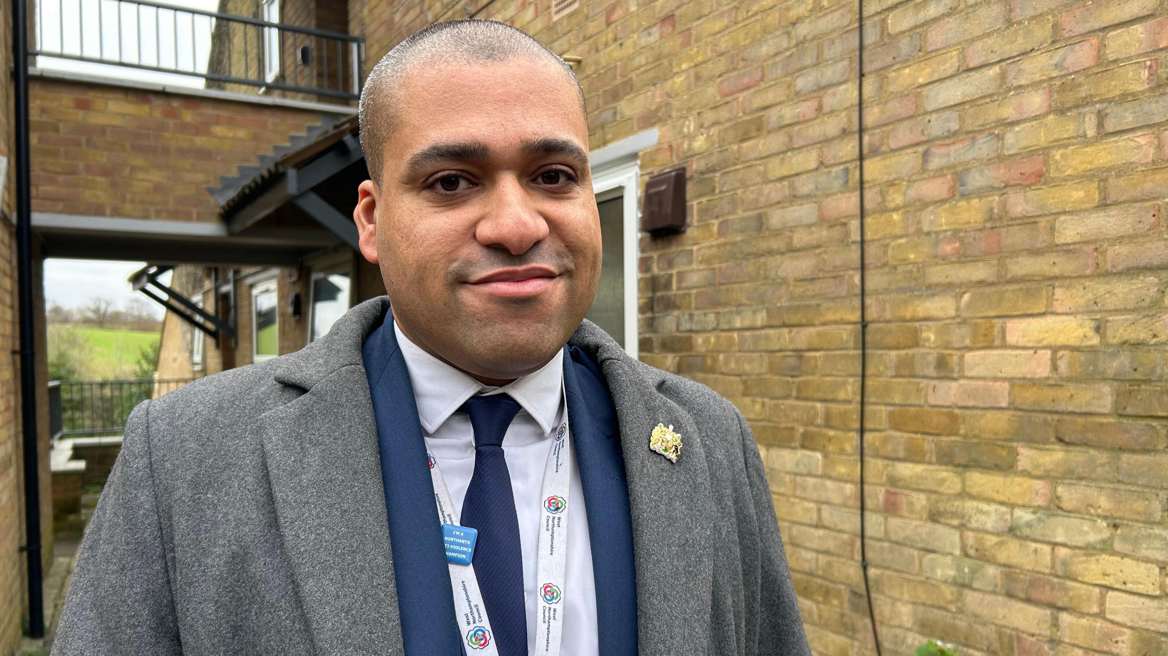 A smiling James Hill looks directly at the camera as he is photographed in front of a block of flats. He is wearing a blue suit and grey coat. 