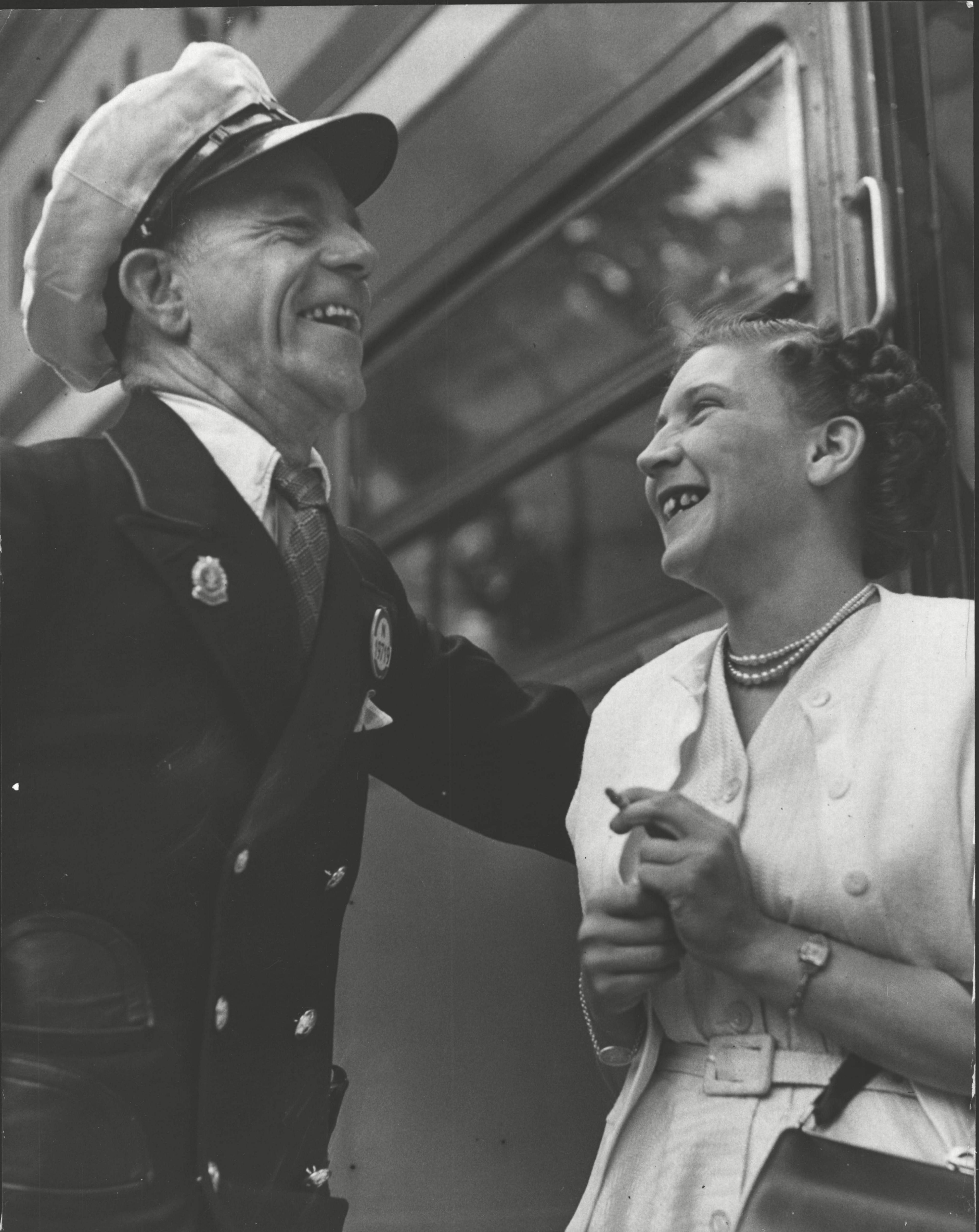 Albert Gunter and May Walshaw facing each other laughing and standing by a bus. Mr Gunter is wearing his bus uniform and is on the left of the picture, looking right and down. Miss Walshaw is on the right and is looking left and up. Miss Walshaw is wearing a pale dress with a matching short jacket and a double string of pearls. She has a handbag over her left elbow and holds a cigarette in her left hand.