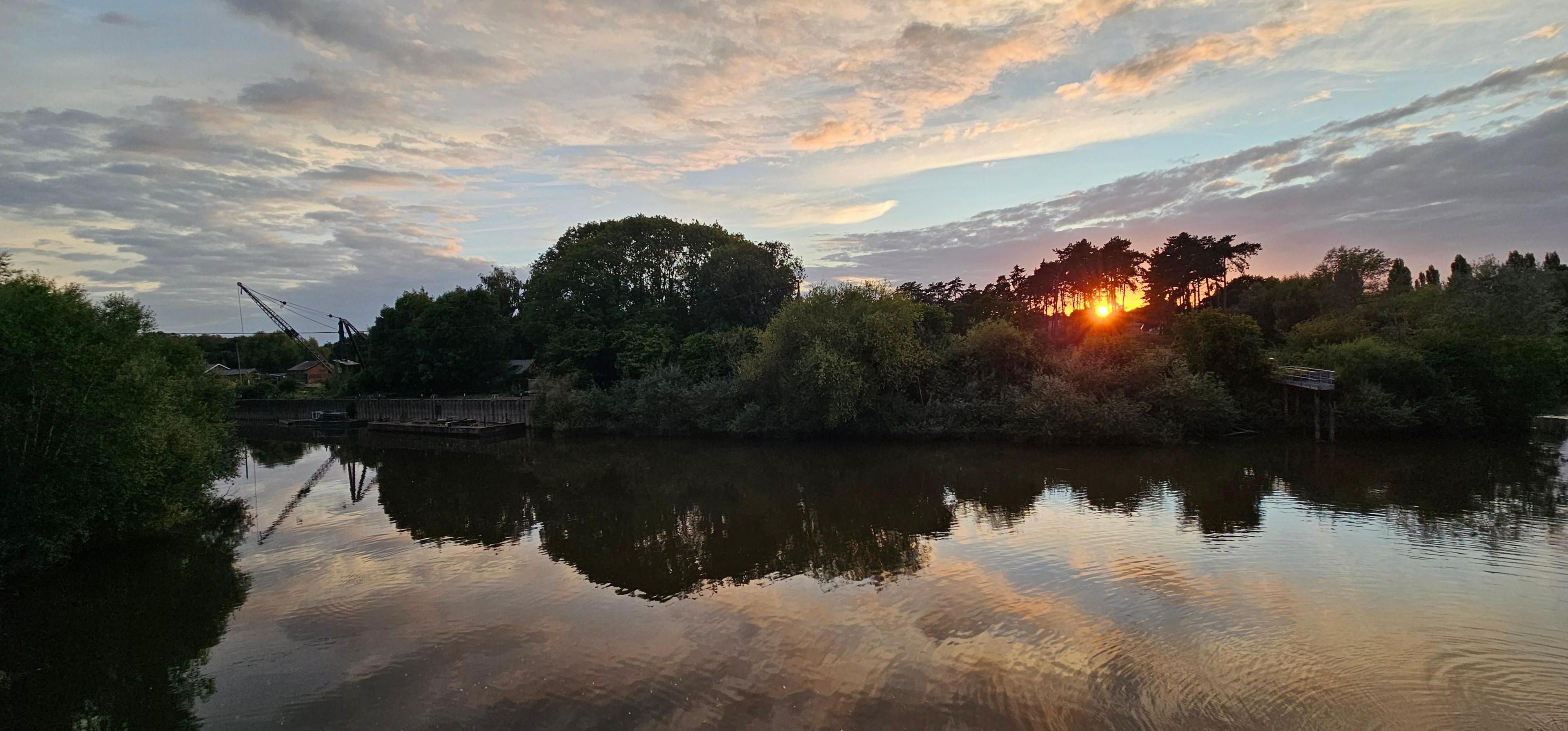 A panoramic shot along the river, showing the trees and their reflection in the water, with the sun going down between the trees. To the left of the shot a crane and its reflection are seen, nearly in silhouette.