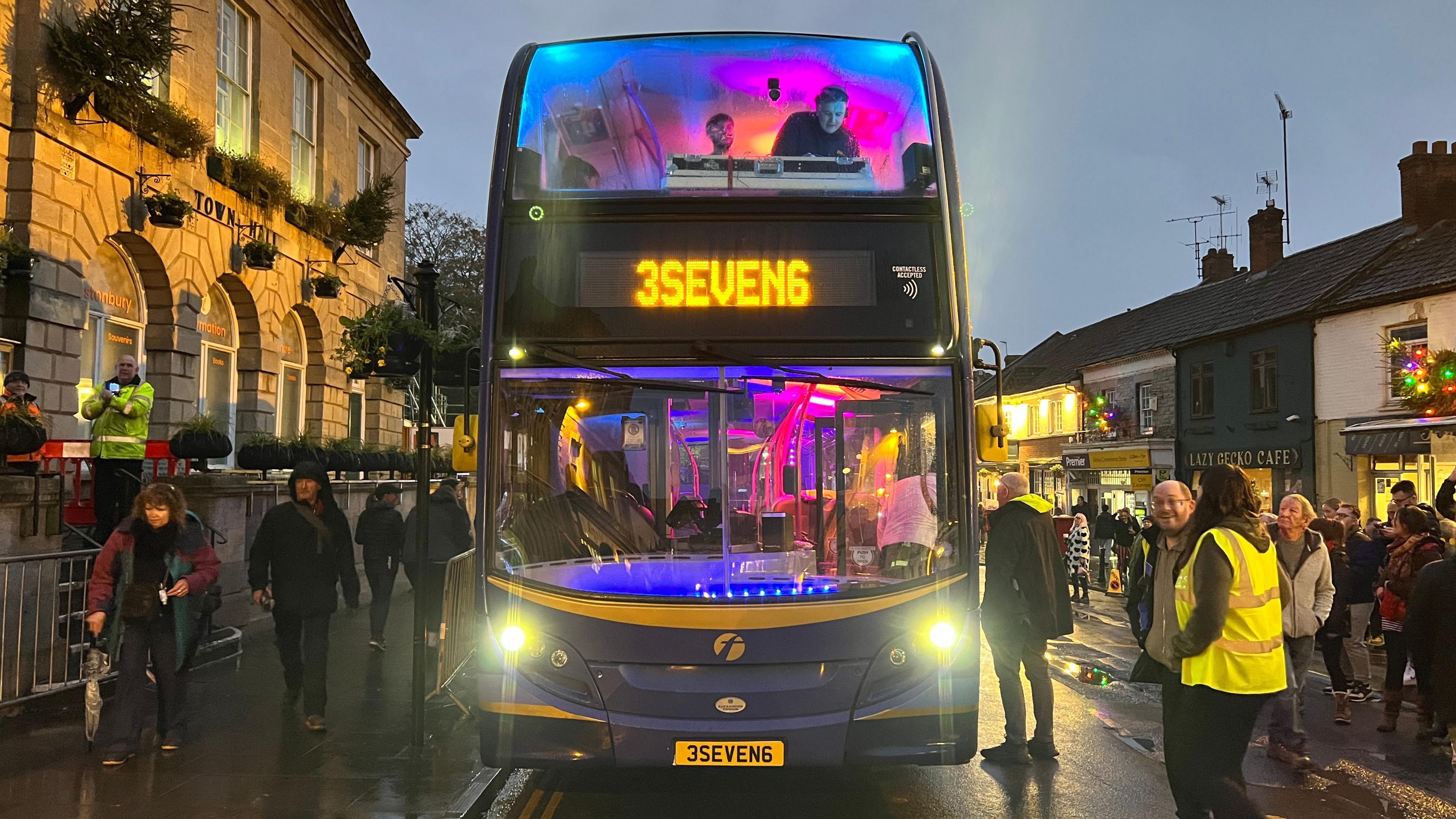 The 376 double decker bus, lit up in pink and purple, at dusk. It is driving down a road in Glastonbury. On the top deck, a member of the 3Seven6 DJ collective can be seen next to a DJ deck. People are lining the street on either side. 