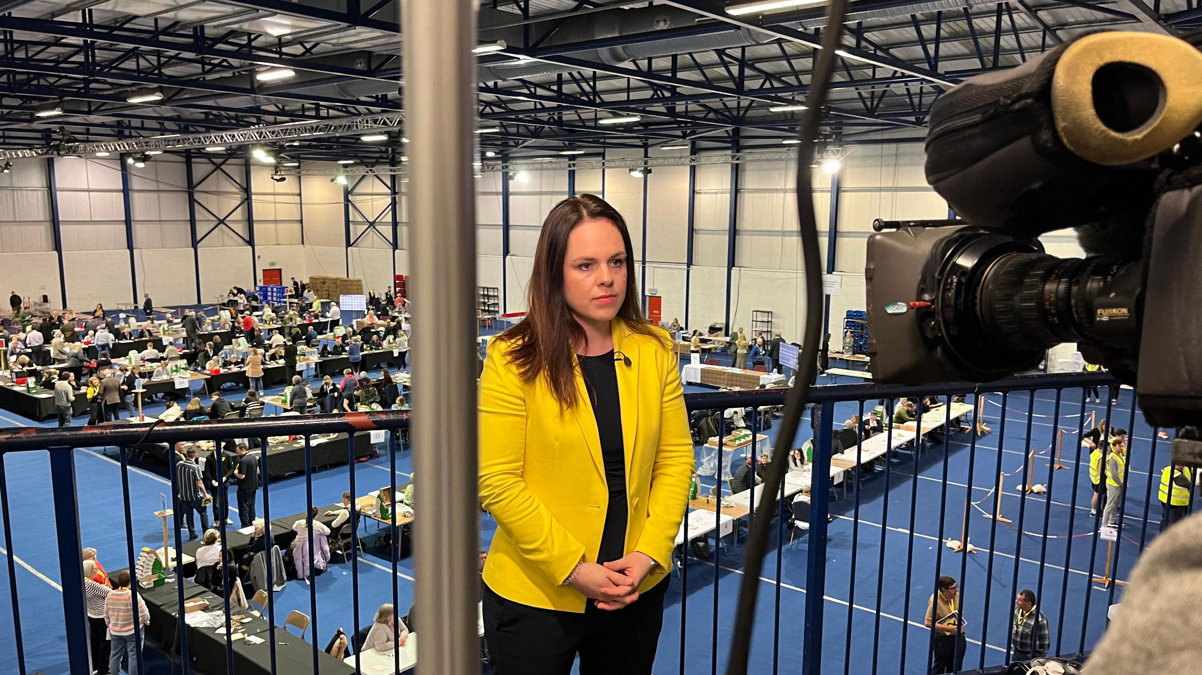 Kate Forbes, in a bright yellow blazer stands on a gantry in front of a TV camera, an electoral count going on at tables in the room below her.