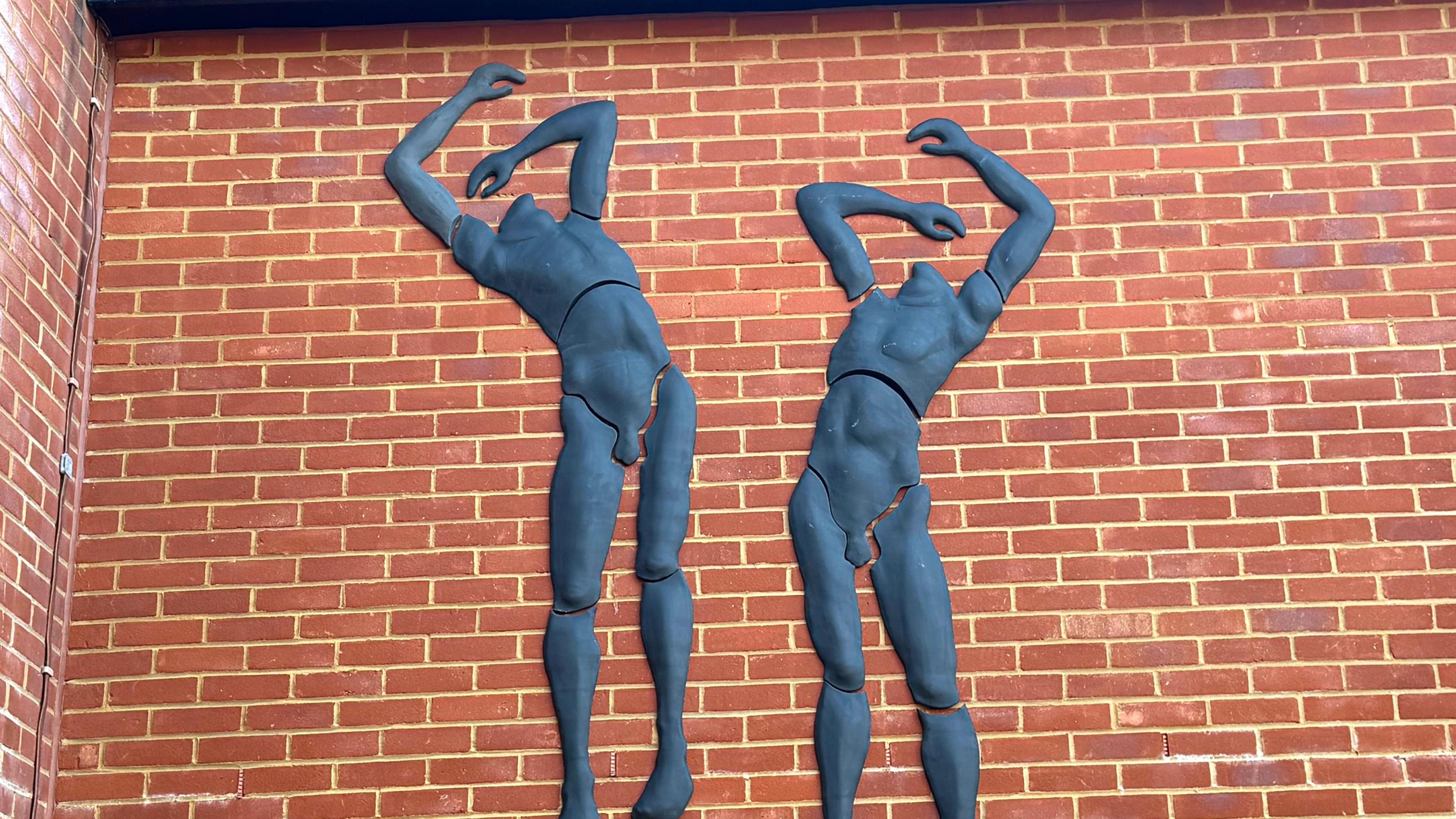 A sculptural artwork attached to a brick wall. The dark figures are shown leaning backwards, as if having been struck, with their aims flailing.