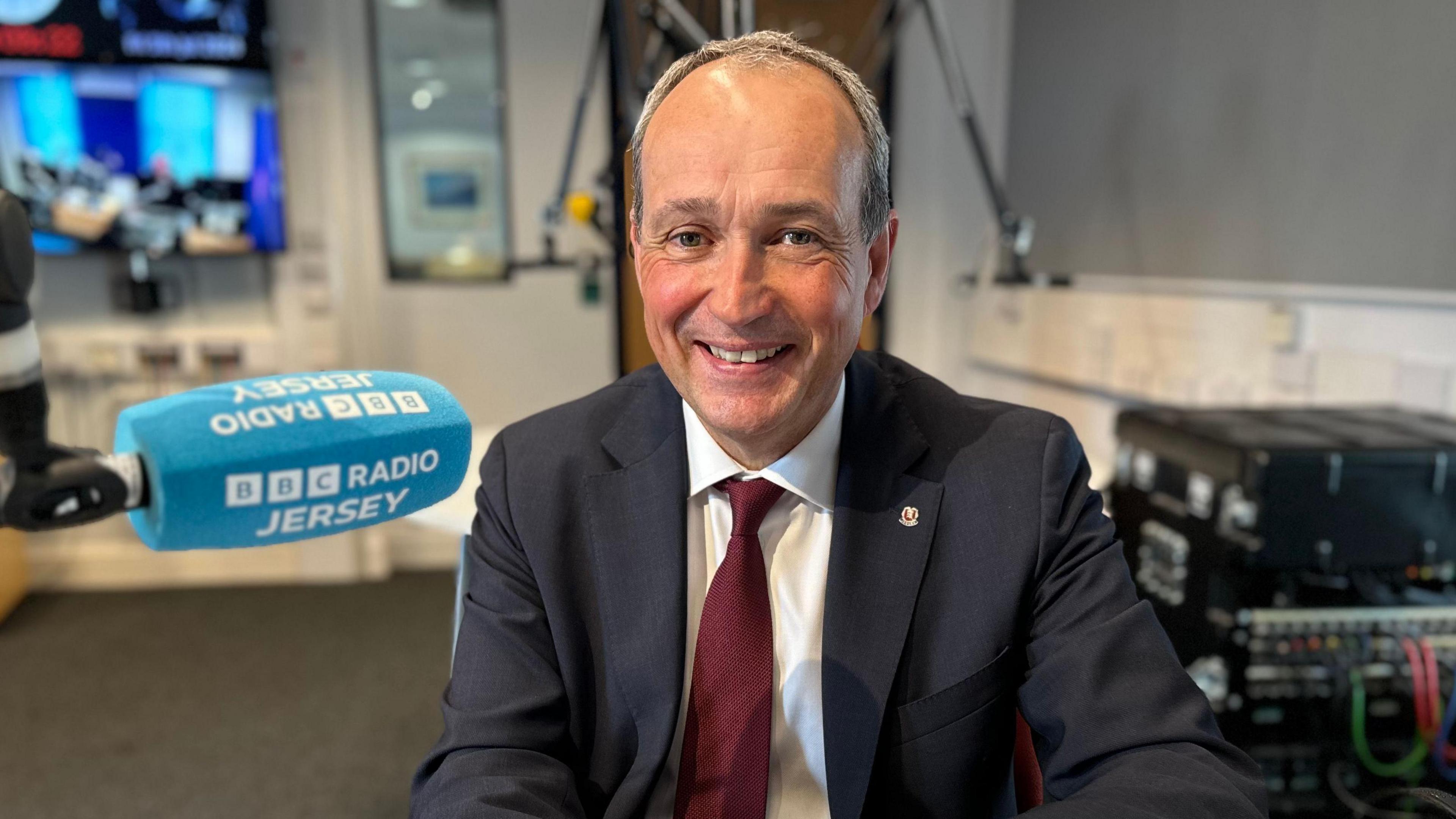 Chief Minister Deputy Lyndon Farnham - a man with grey hair, centrally bald, large grin, wearing a black suit, a white shirt and a red tie, he is in a BBC Radio Jersey studio