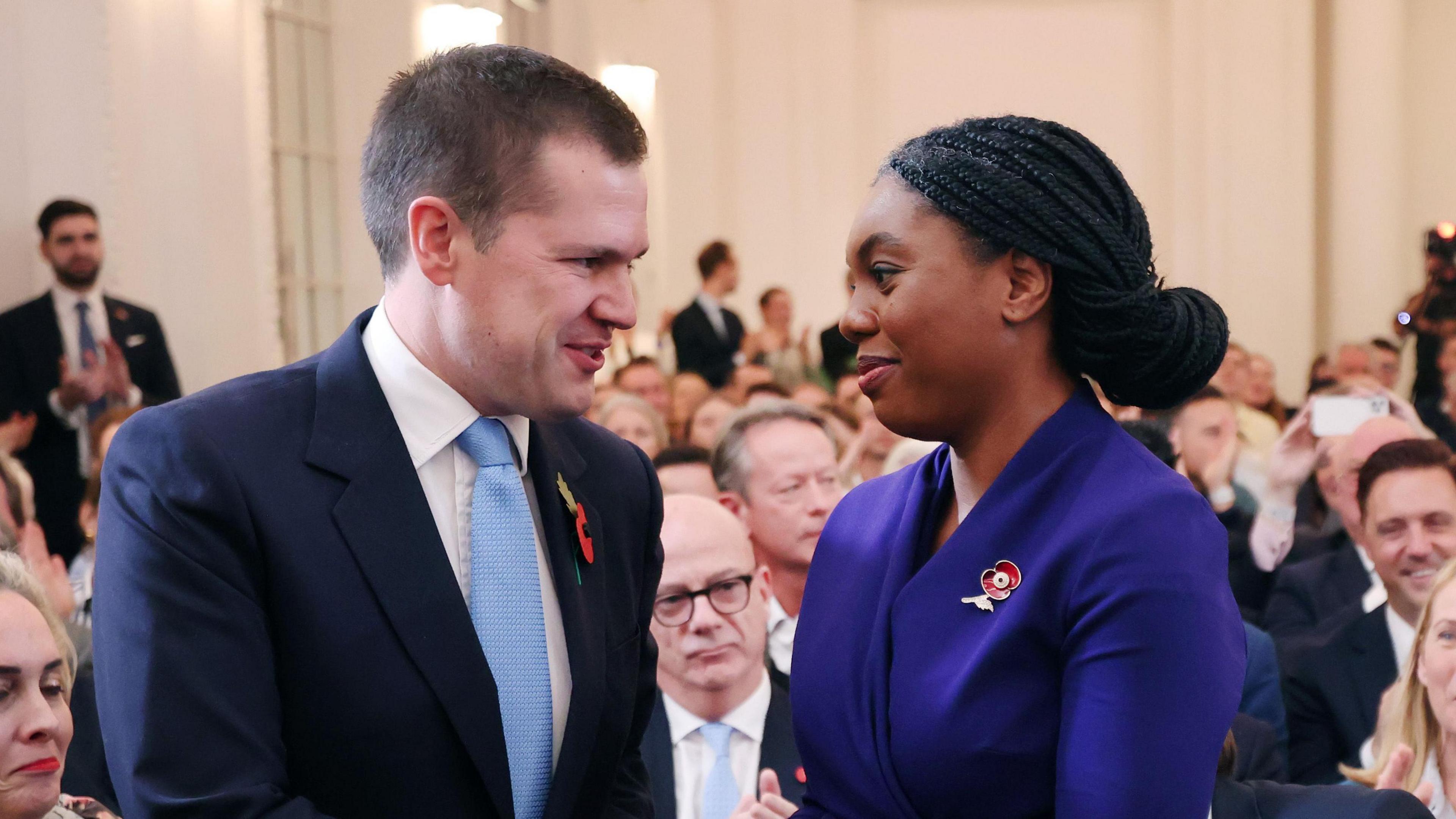 Robert Jenrick (left) congratulates Kemi Badenoch on her success as the results of the leadership election are announced. Conservative Party members appear to have mixed reactions to the announcement in the background.