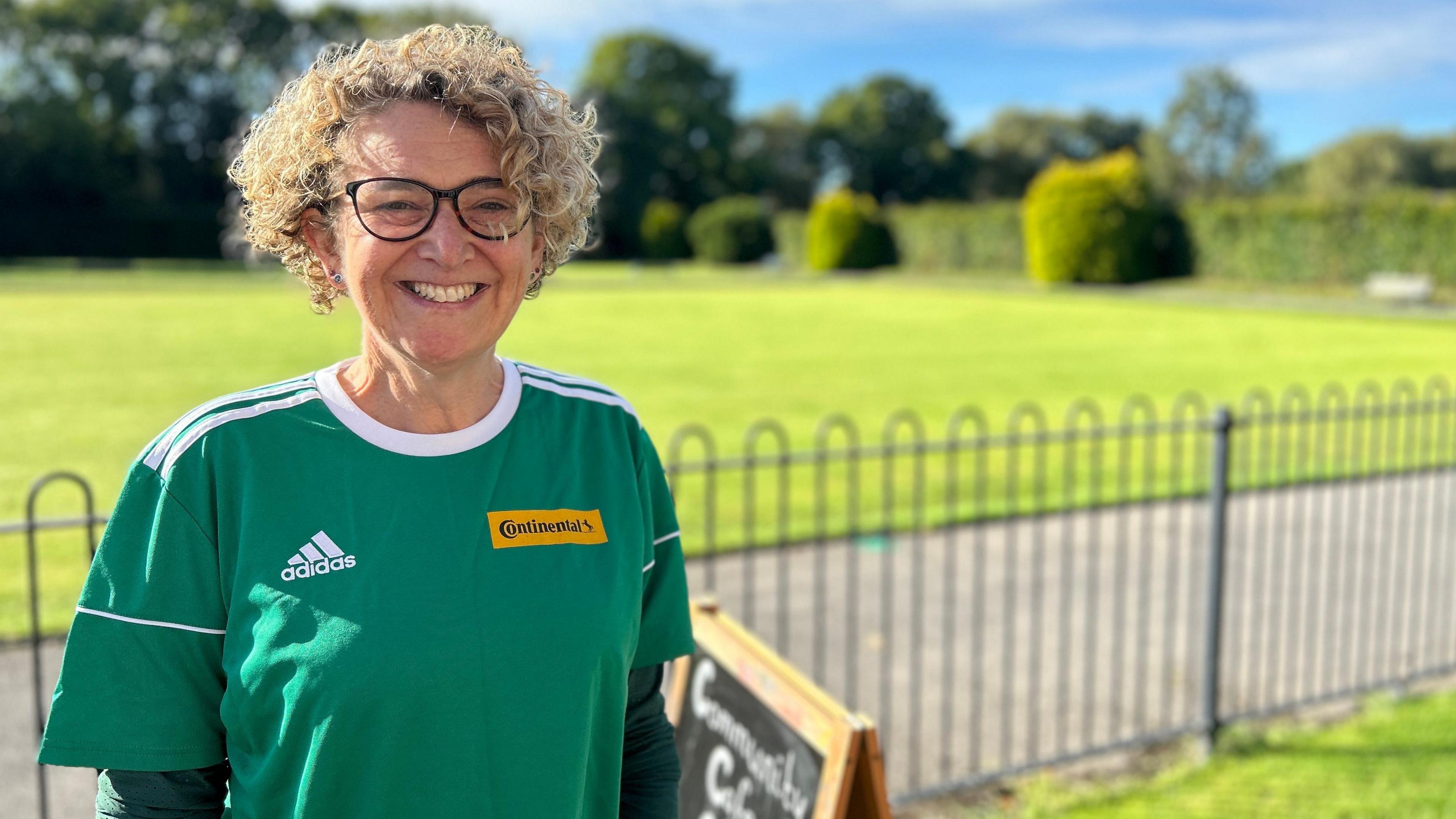 Jane is stood in front of a park scene with a sports shirt and a big smile on her face