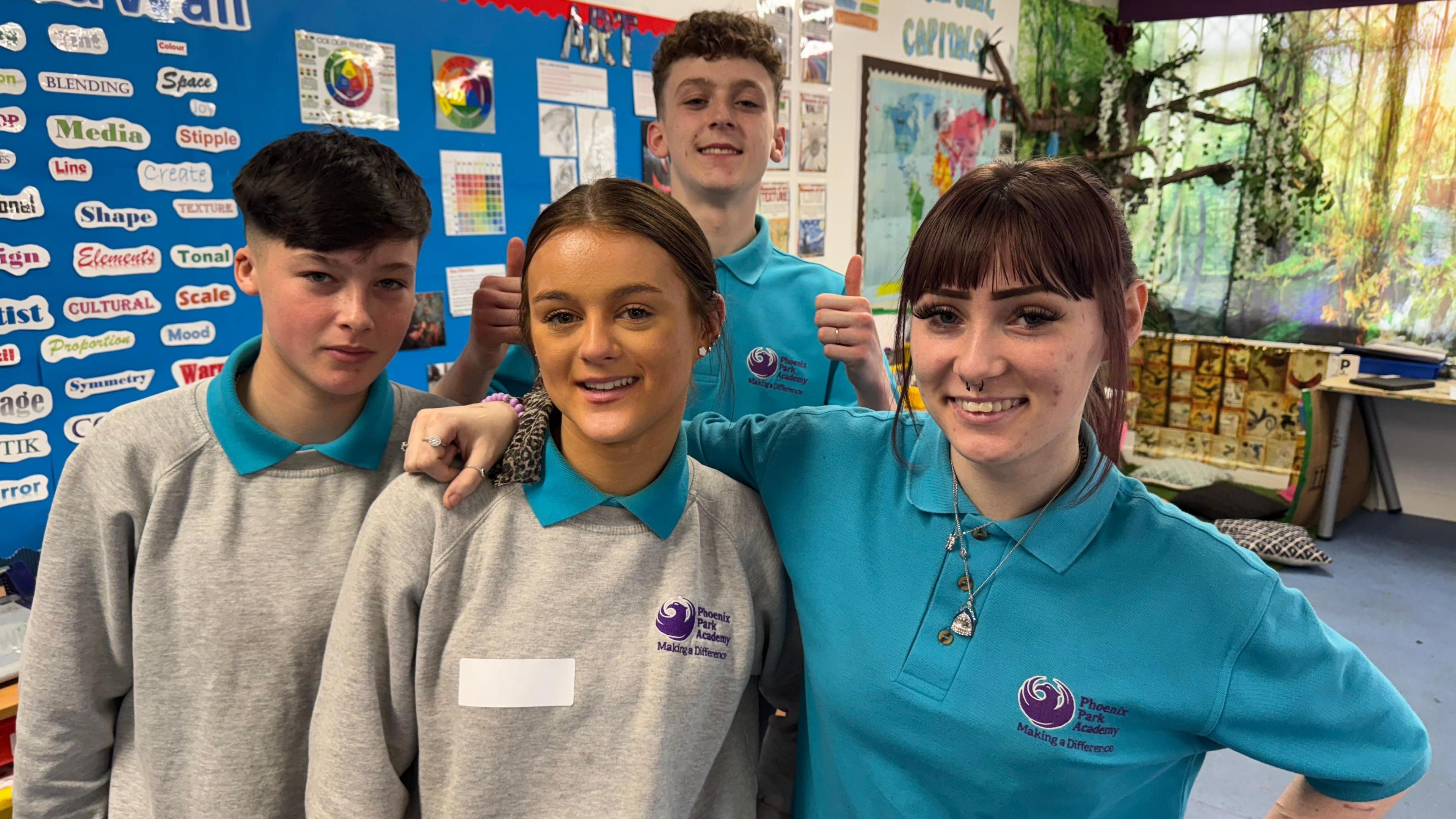 Four young students in a classroom. Two boys and two girls. One of the boys is stood behind the other three with both thumbs up, while one girl has her arm round the other. All four are posing for the camera.