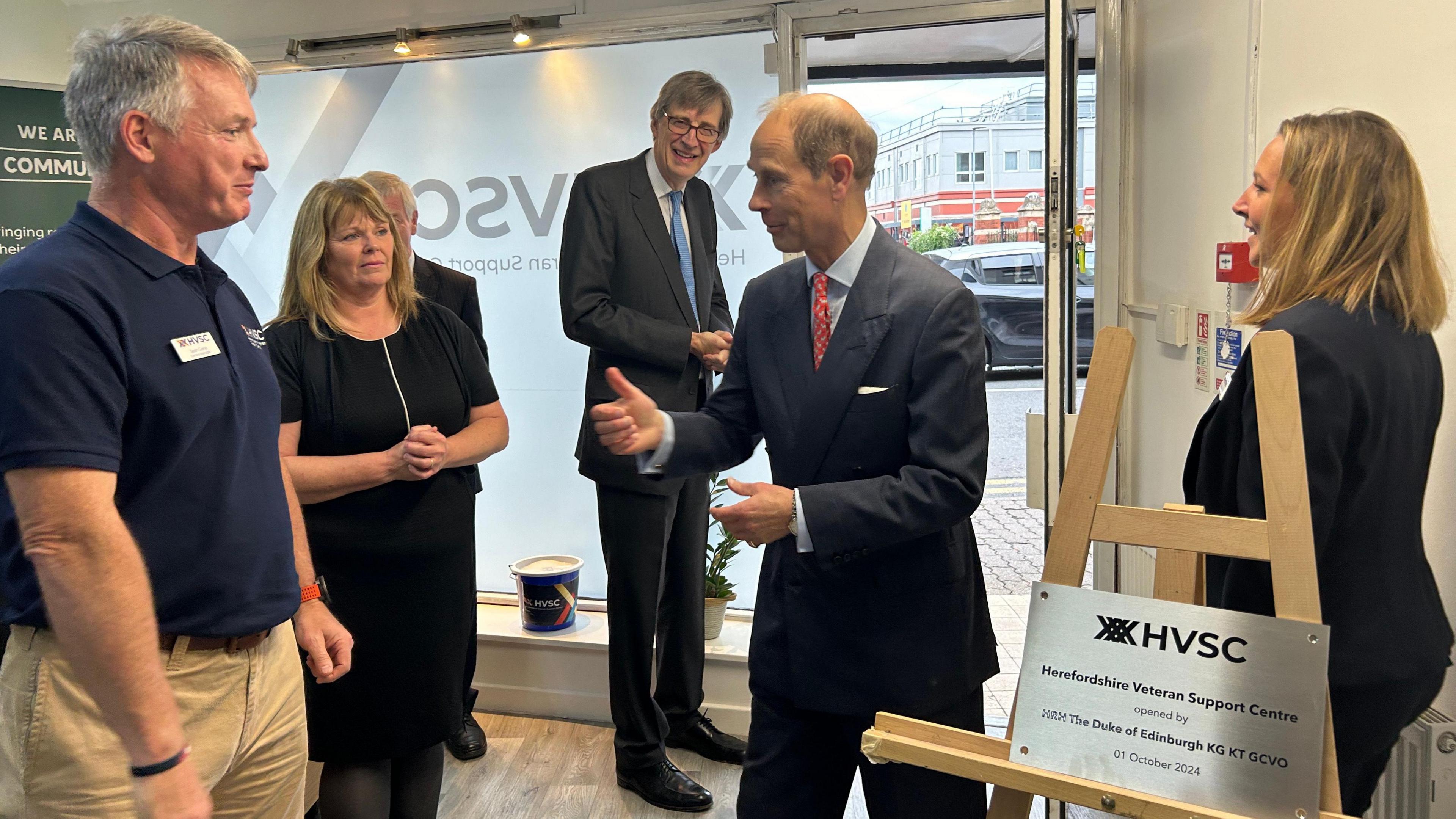 The Duke of Edinburgh speaking to a room of five staff members. He is dressed in a suit with a red tie. 