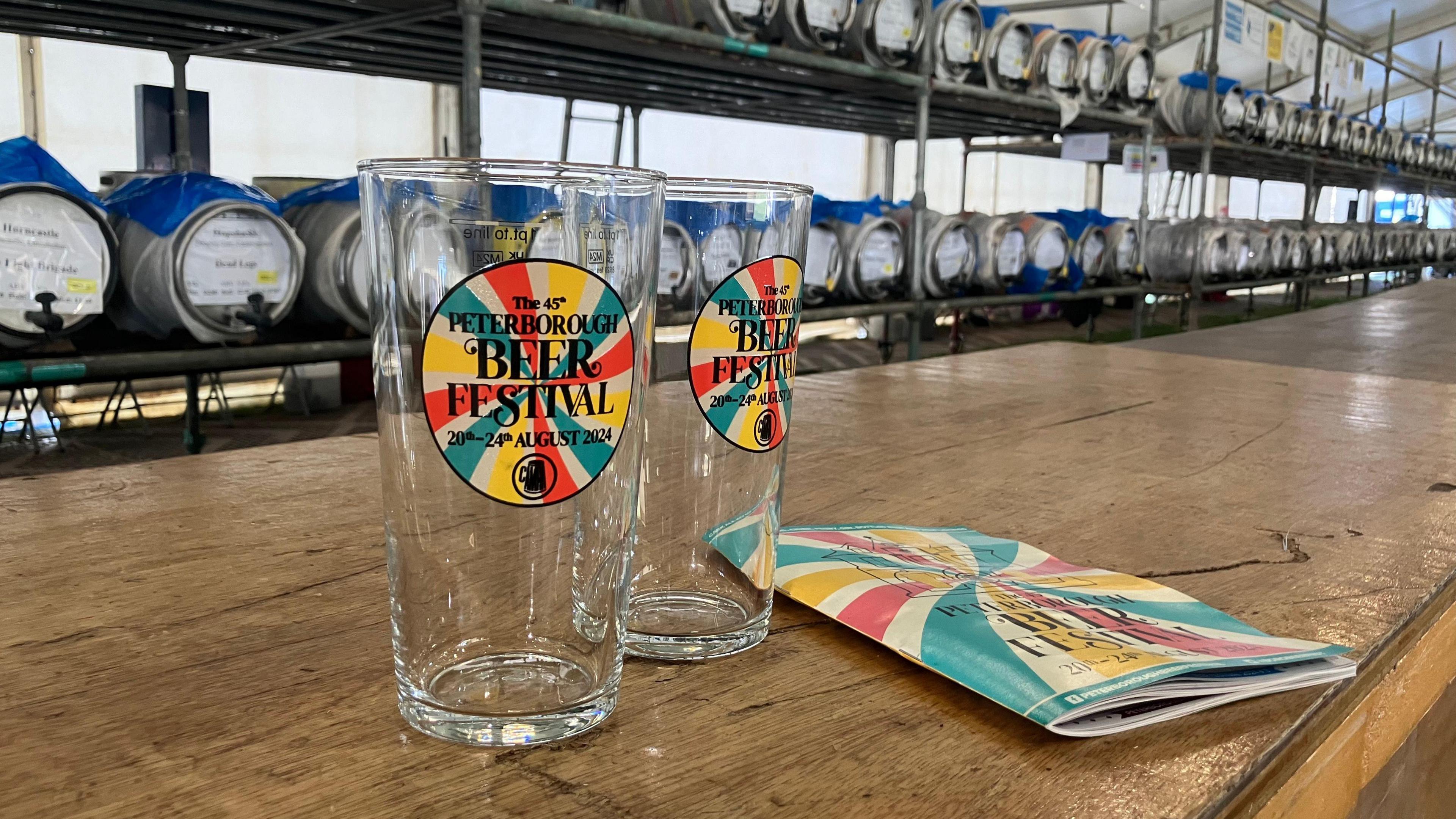 Empty glasses with "beer festival" written on them standing on a wooden bar, with rows of barrels behind them