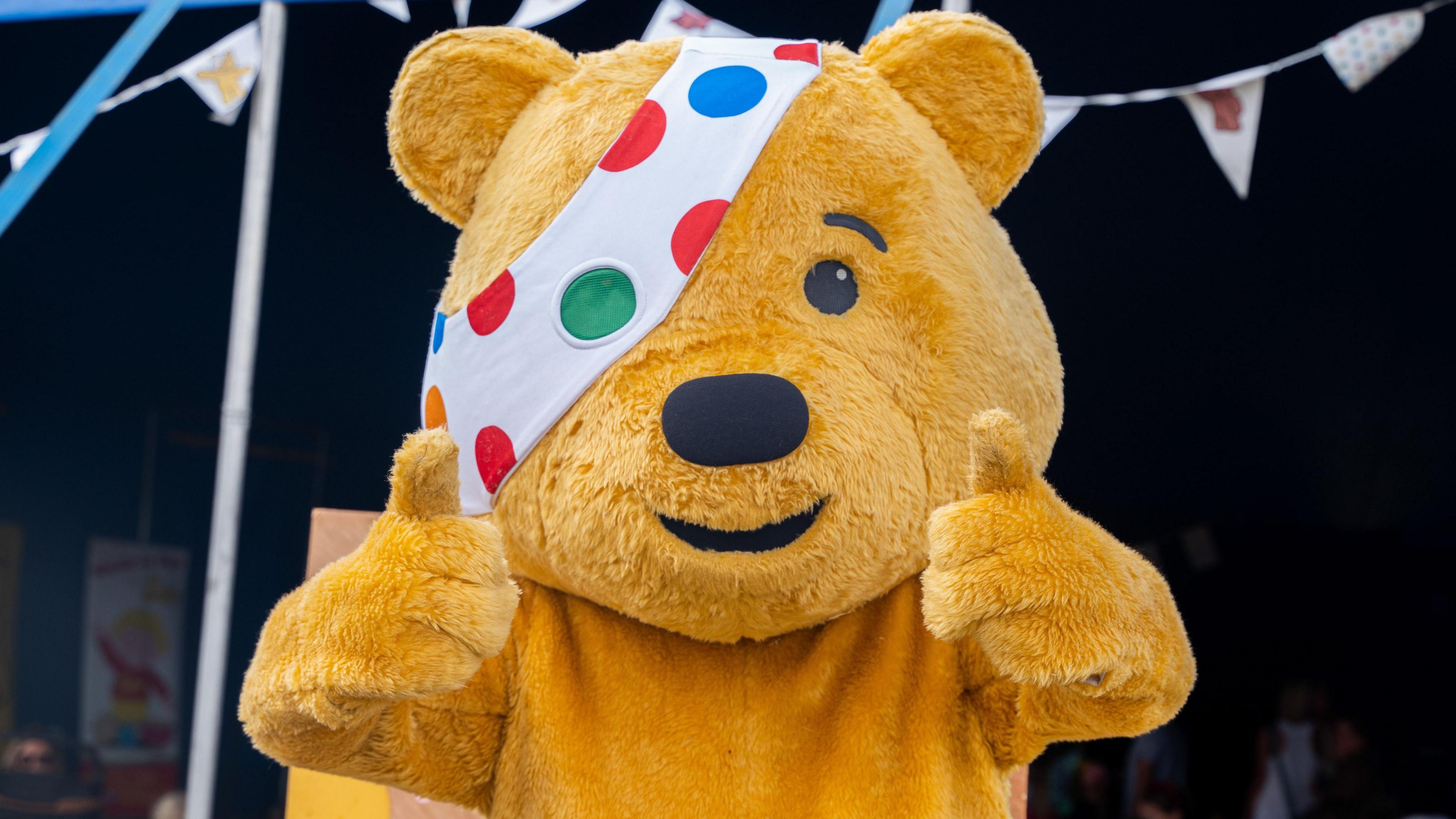 A yellow children in need bear with a white, spotty eye patch is looking directly at the camera with his thumbs up