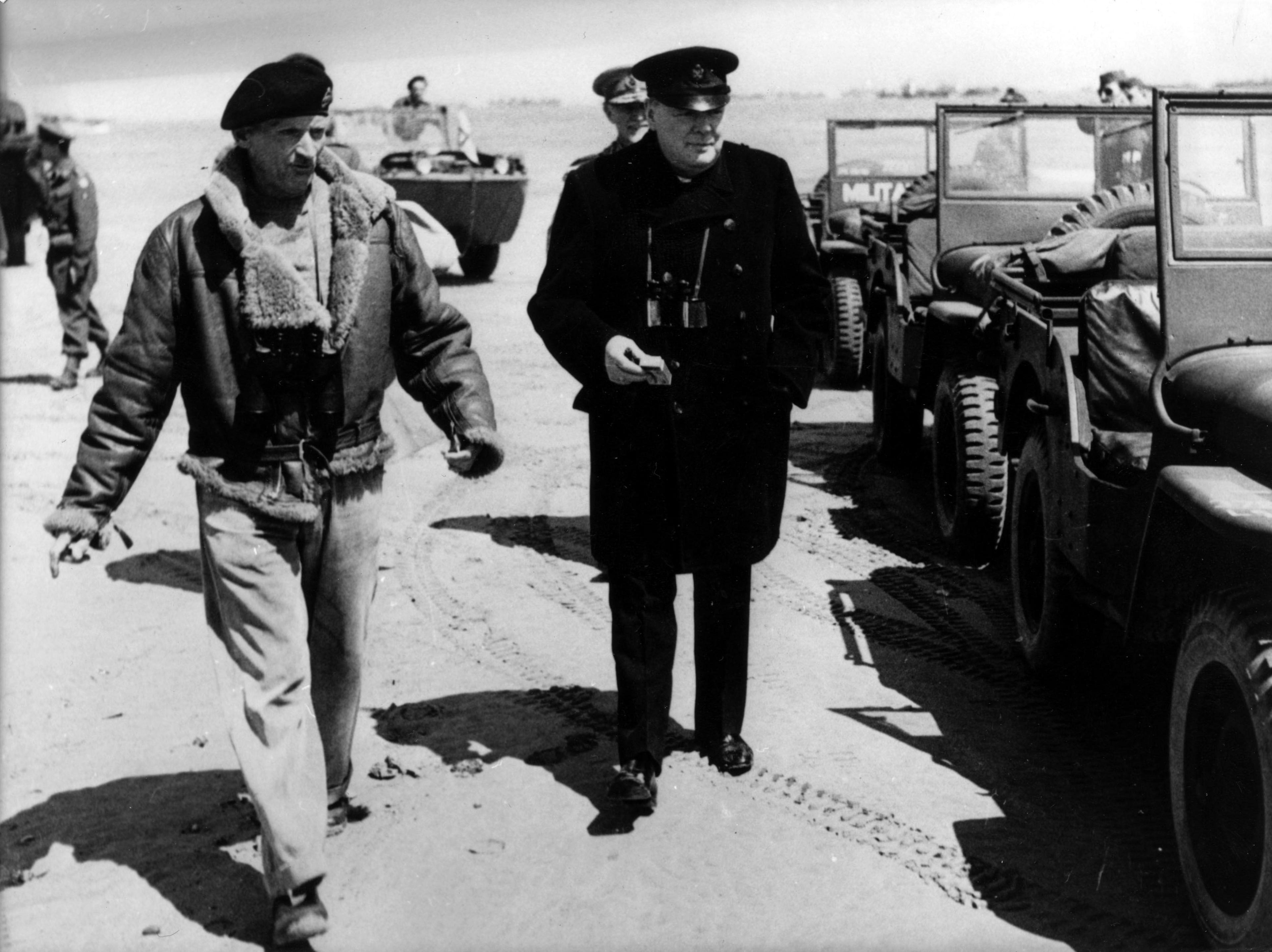 Sir Winston Churchill stands next to a row of military jeeps on a beach in Normandy. He is wearing a black overcoat, trousers and military hat. He is alongside Field Marshall Bernard Montgomery, who is wearing a beret and fur coat.