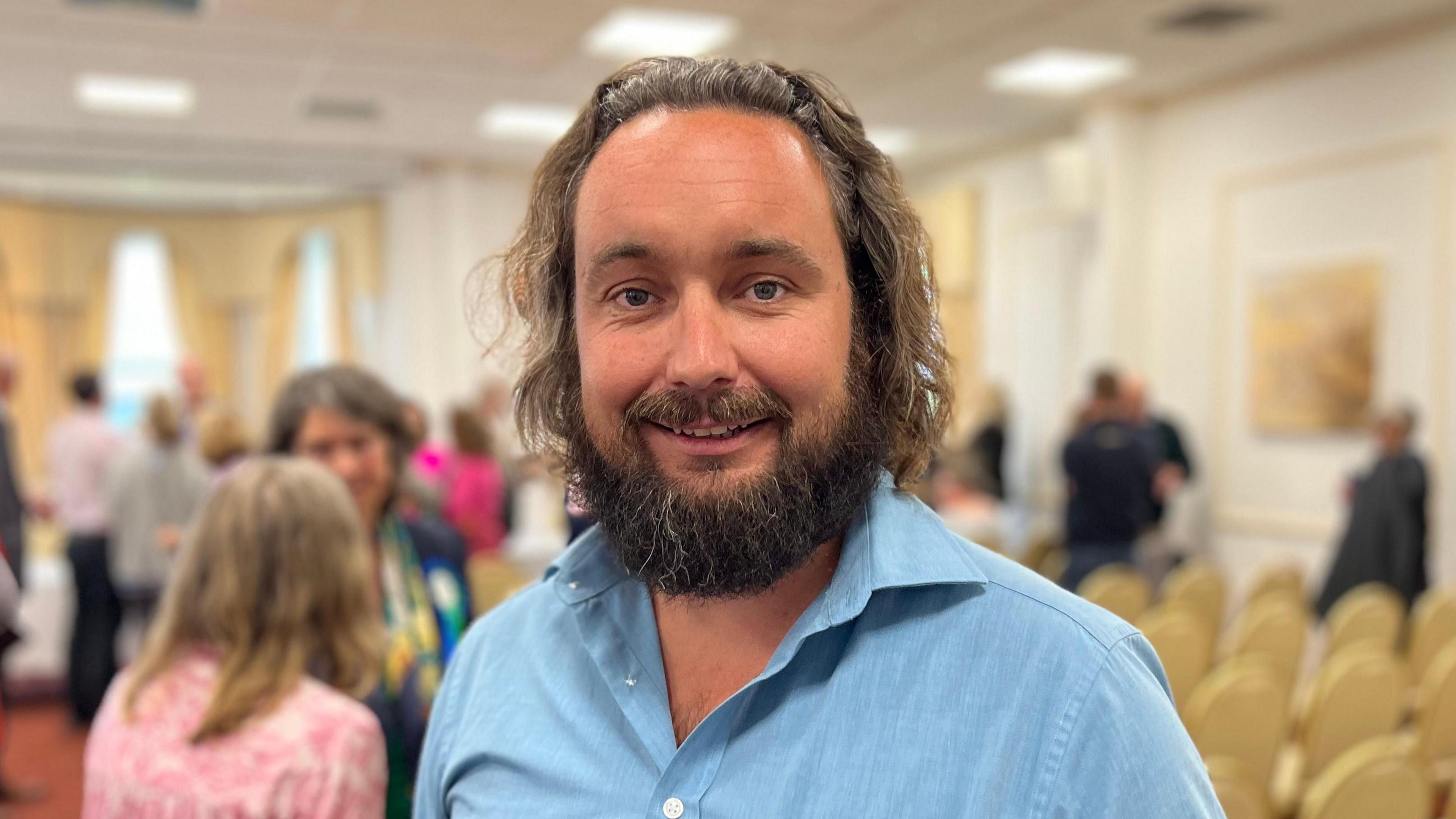 Deputy Lee Van Katwyk - A man with long brown hair, and a big bushy brown moustache and beard. He is wearing a blue shirt buttoned down to show some of his chest. 