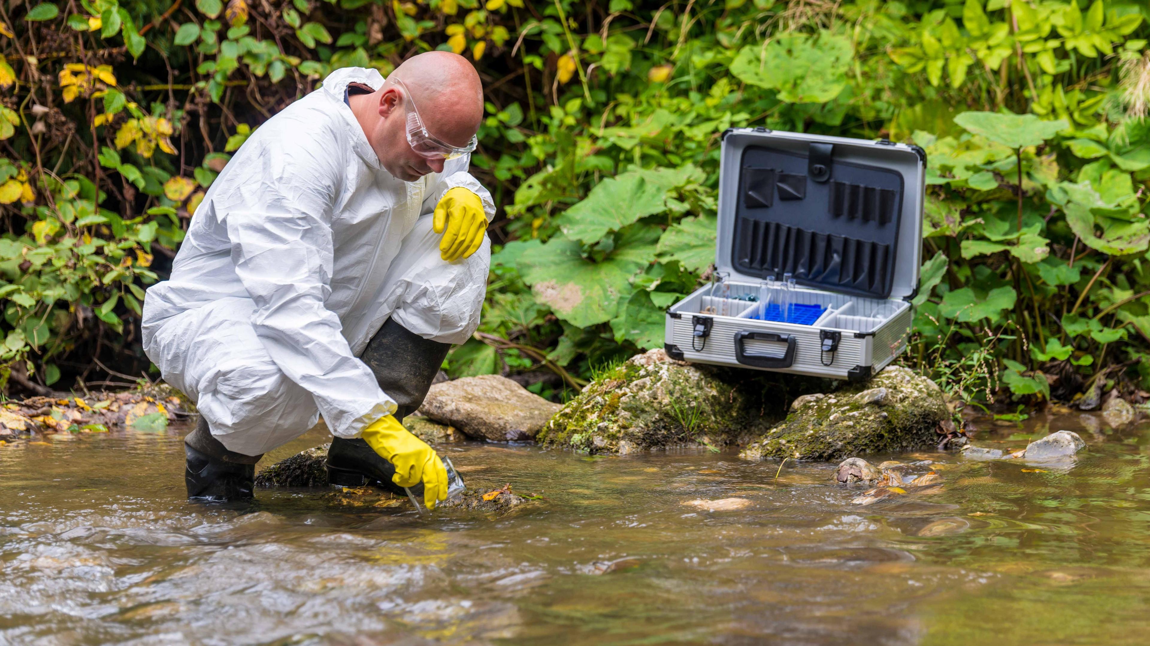 General image of a scientist in a white waterproof suit, with black boots and yellow gloves, testing the river water for pollution